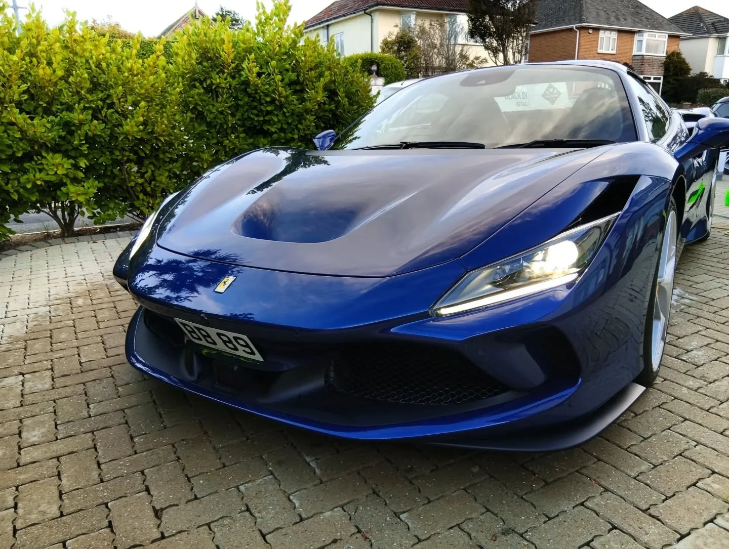 A blue Ferrari sports car parked on a brick driveway with green shrubbery and houses in the background.