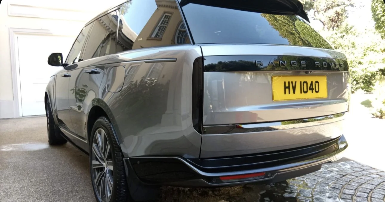 A silver Range Rover parked on a driveway with a yellow license plate HV 1040. The reflection of a house and trees can be seen on the rear window and body of the vehicle.