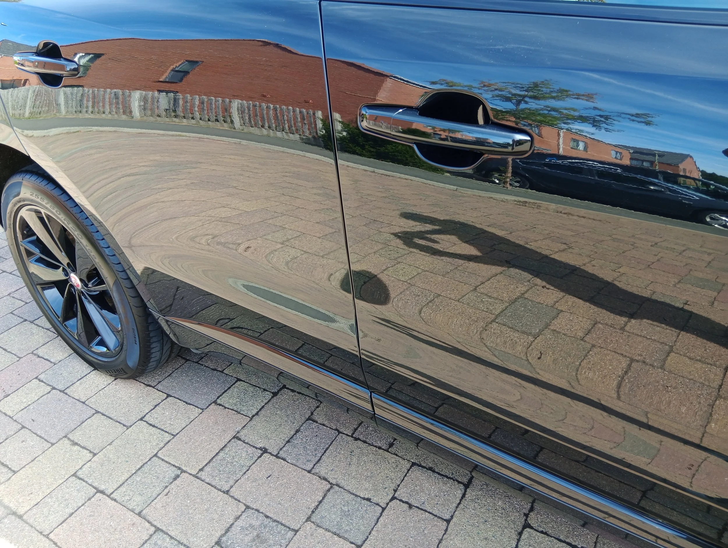 Close-up of a black car's side, showing reflection of houses and trees on its shiny surface, parked on a paved driveway.
