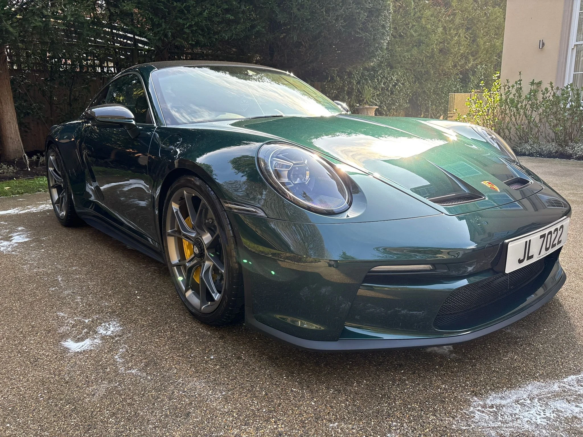 A green Porsche sports car parked on a driveway with trees and a house in the background.