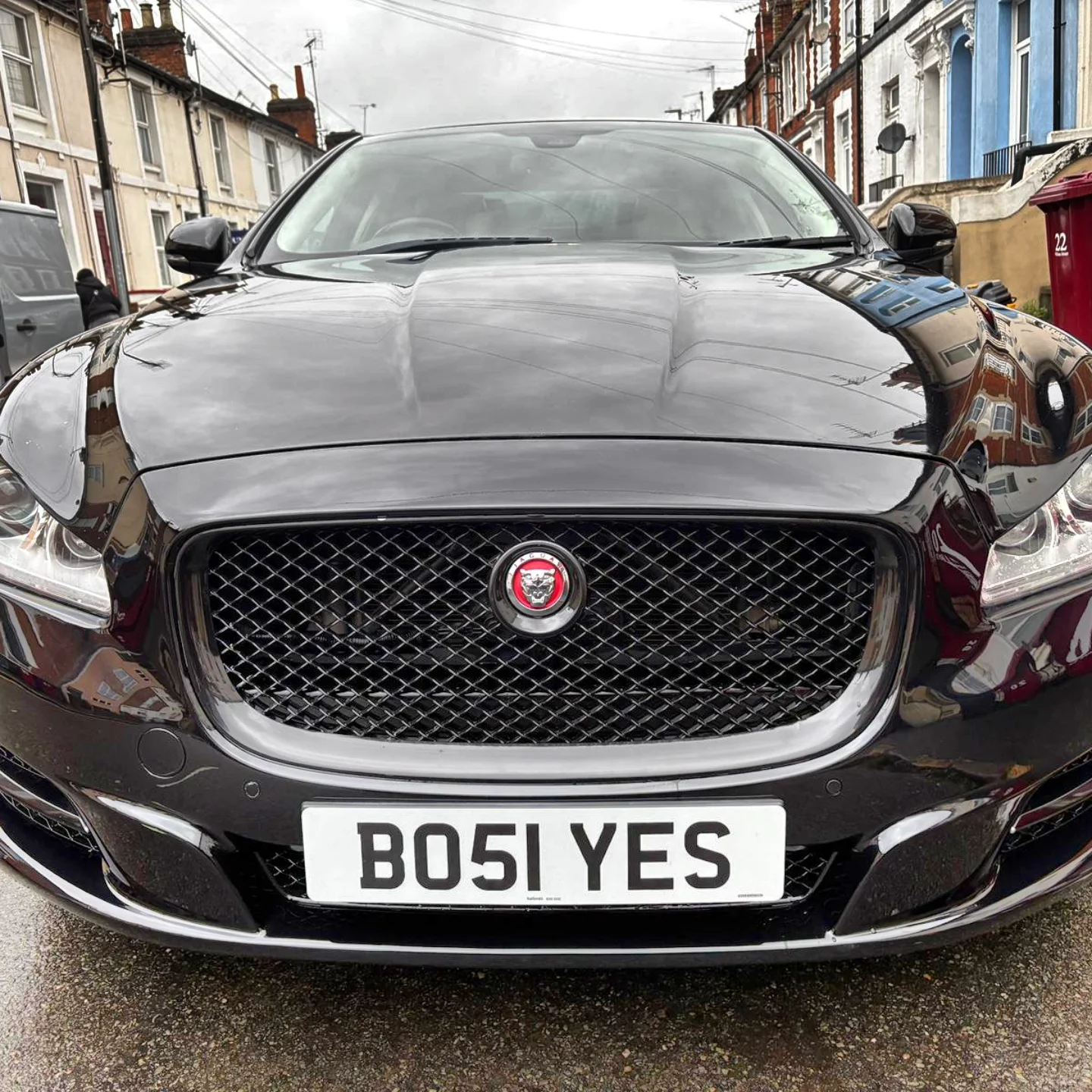 Black Jaguar XF parked on the street with reflective surface showing neighborhood houses. The car has a custom license plate reading 'BO5 I YES' and a Jaguar emblem on the grille.