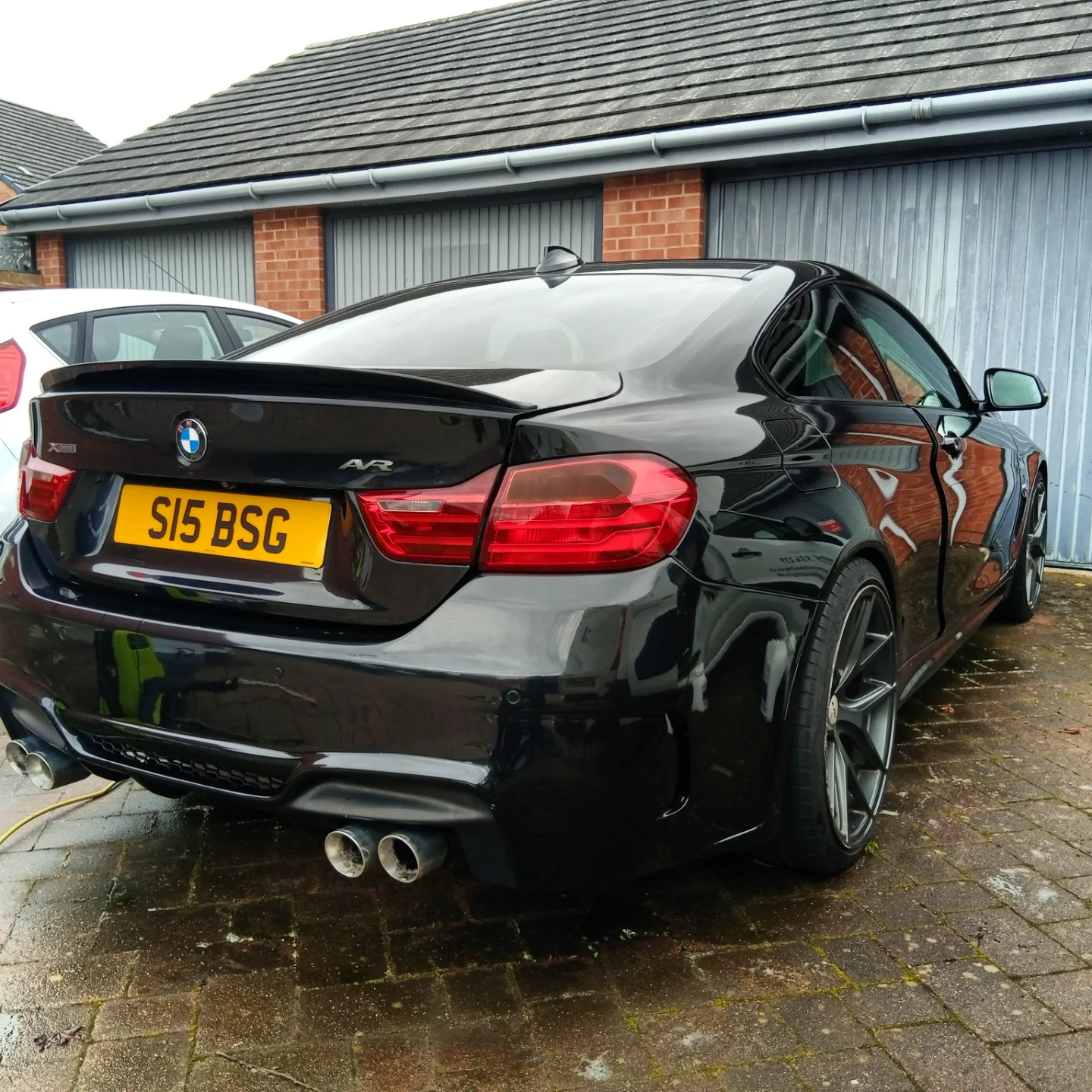 Black BMW M3 sports car parked on brick driveway near house with blue garage doors.