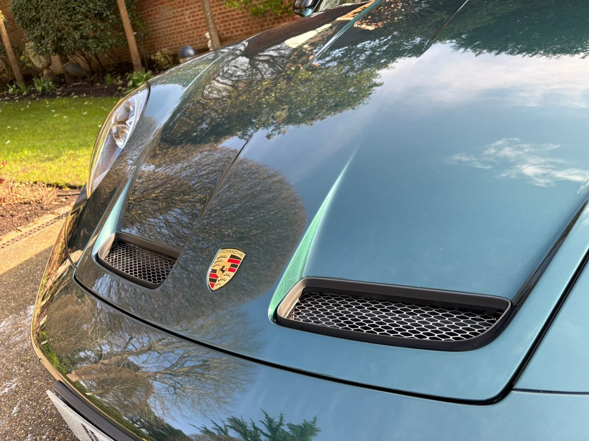 Close-up of the front part of a black Porsche sports car, showing the front grille and part of the hood with the Porsche emblem, parked outdoors with trees and a brick wall reflected on its surface.