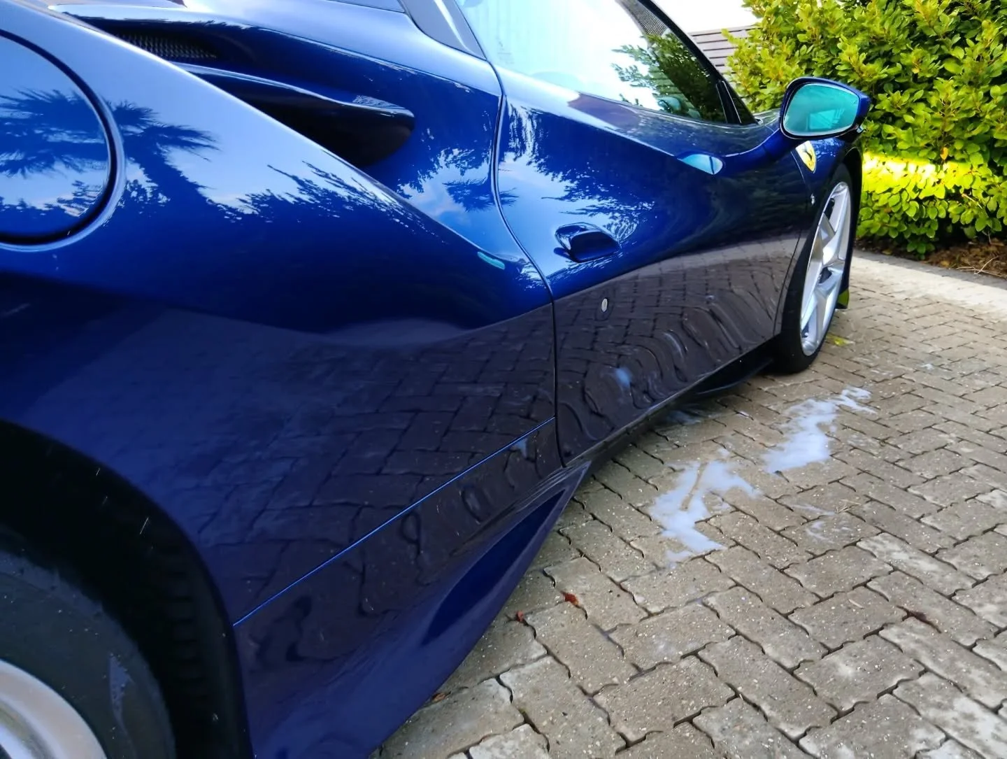 A close-up of a shiny blue sports car parked on a brick driveway, with soap suds on the ground nearby and green bushes in the background.