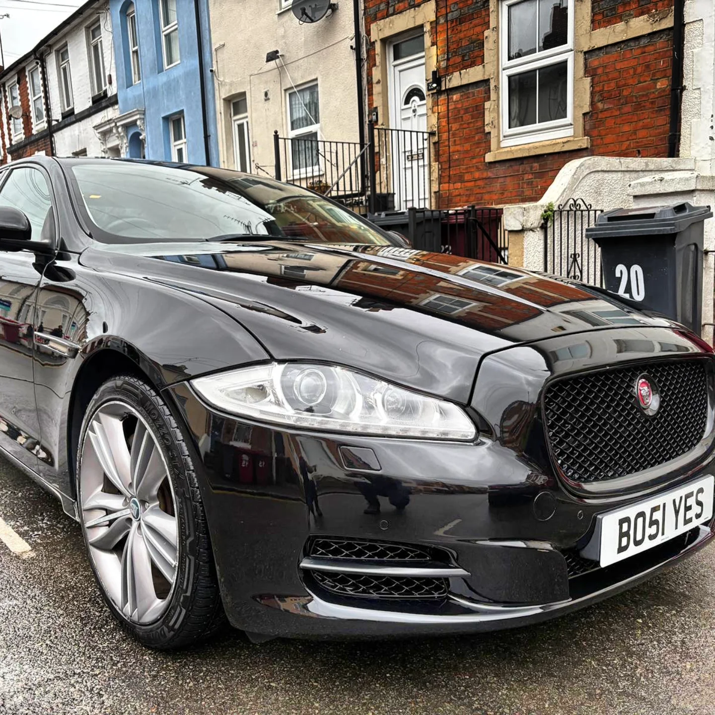 Black Jaguar XE parked on a wet street in front of colorful residential buildings with brick facades, with the front of the car visible and the reflection of buildings on its surface.