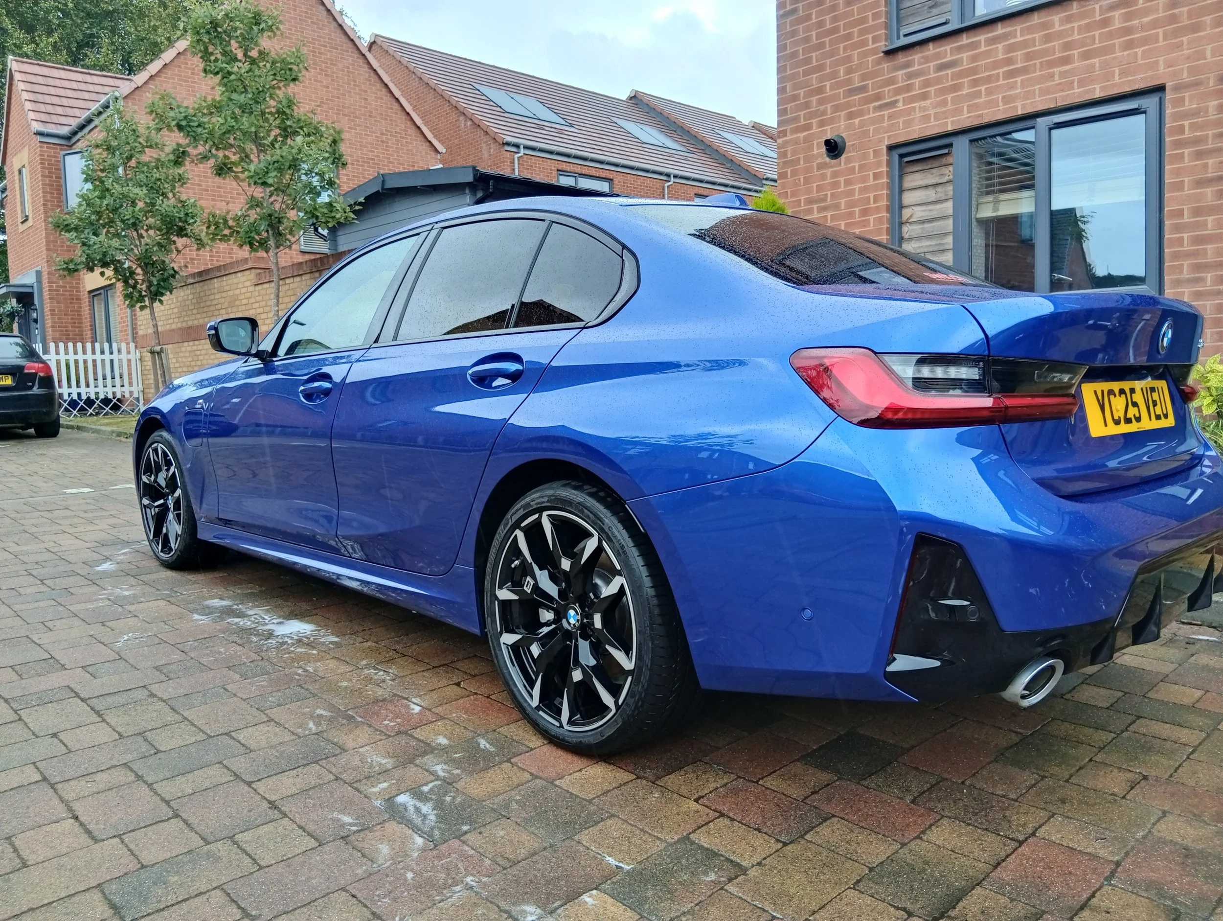 Blue BMW sedan parked on a brick driveway after a rain shower, with wet pavement, residential houses, and a tree in the background.