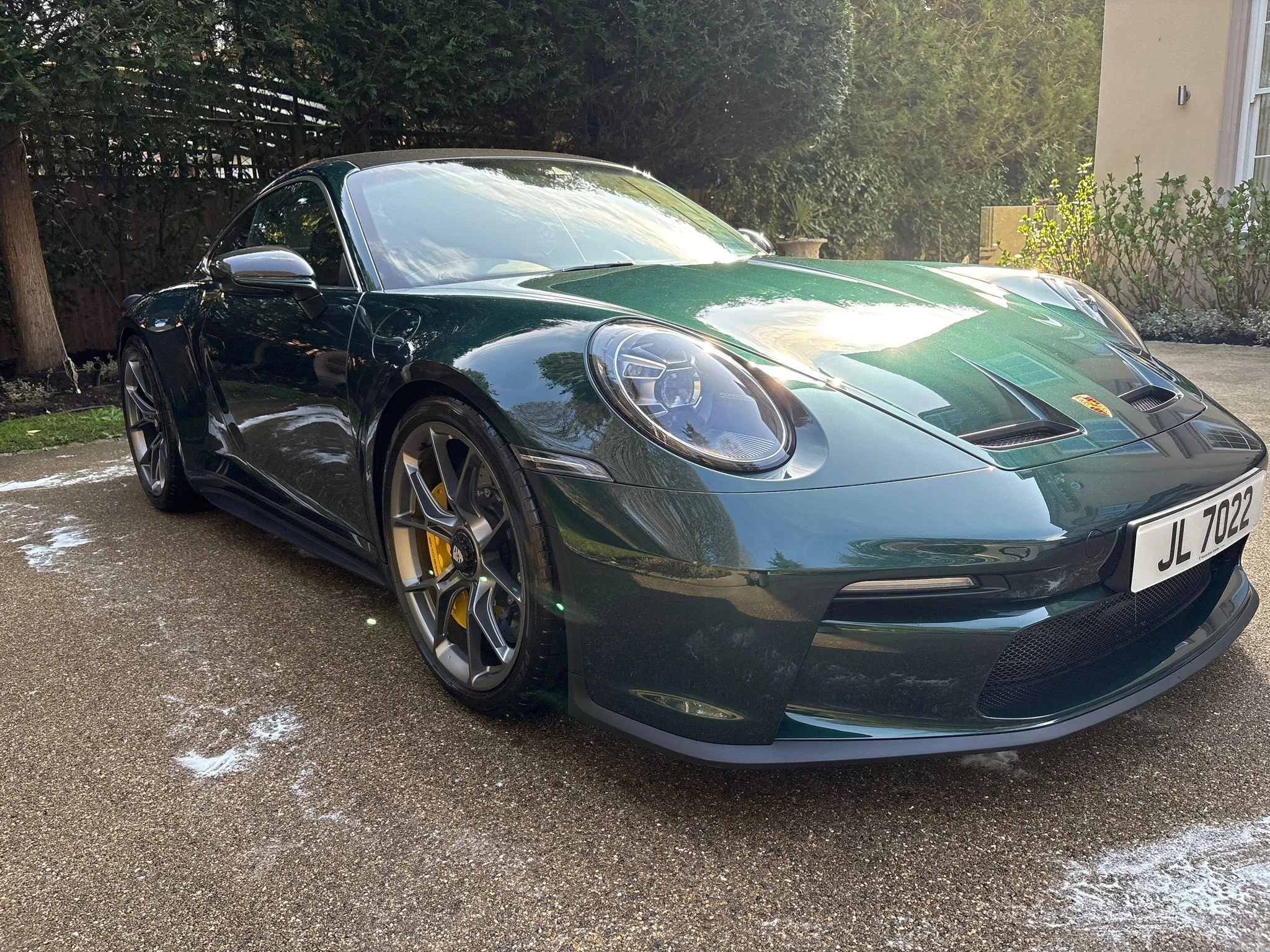 A dark green Porsche sports car parked on a driveway with trees and bushes in the background.