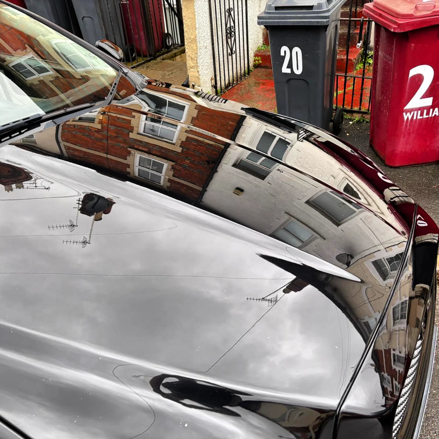 Reflections of residential buildings and a cloudy sky on the shiny hood of a black car parked on a street lined with trash bins and a gate.