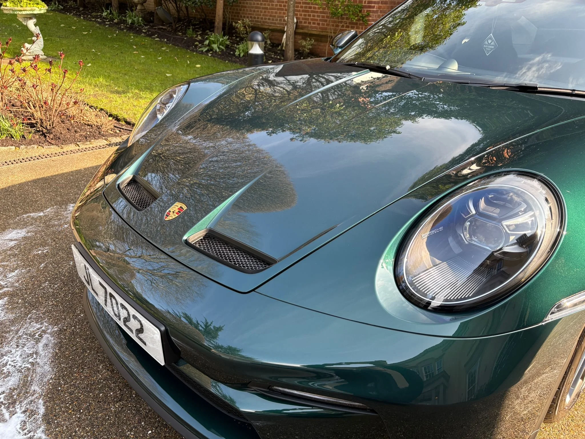 A dark green Porsche sports car parked on a residential driveway, with a garden and a brick wall in the background, and the car's shiny surface reflecting trees and the sky.