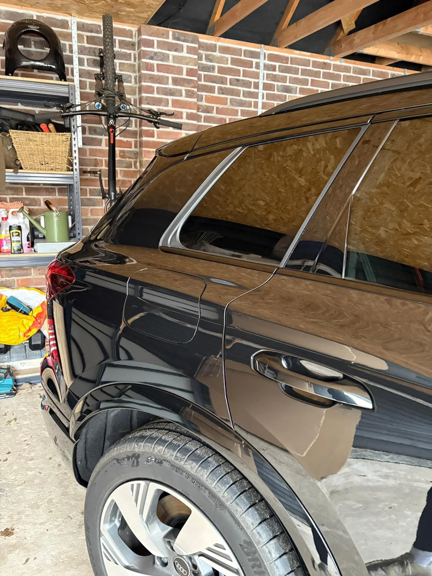 Black SUV parked in a garage with a brick wall and tool shelf in the background. The garage has an unfinished ceiling with exposed wooden beams, and the SUV's surface is reflecting the surroundings.