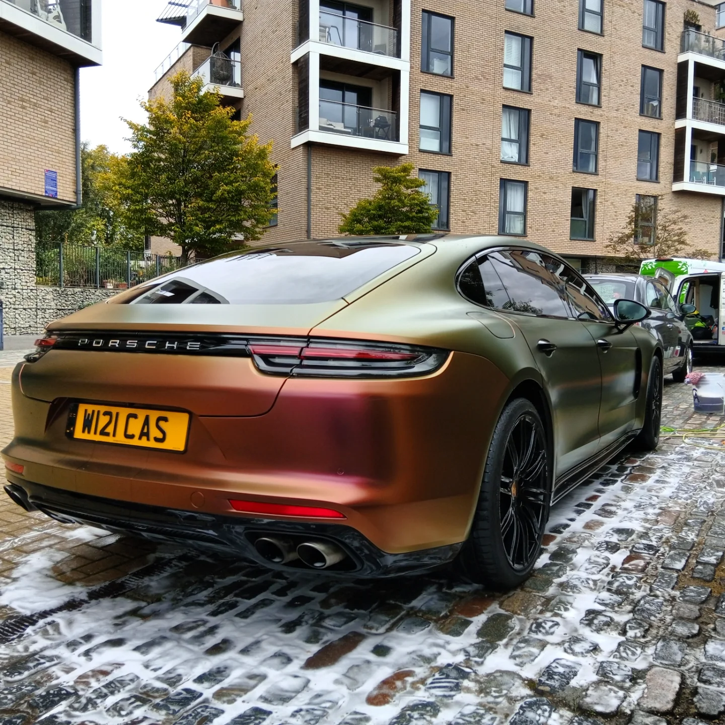A bronze-colored Porsche sports car parked on a cobblestone street with soap suds on the ground, residential apartment buildings in the background, and some trees.