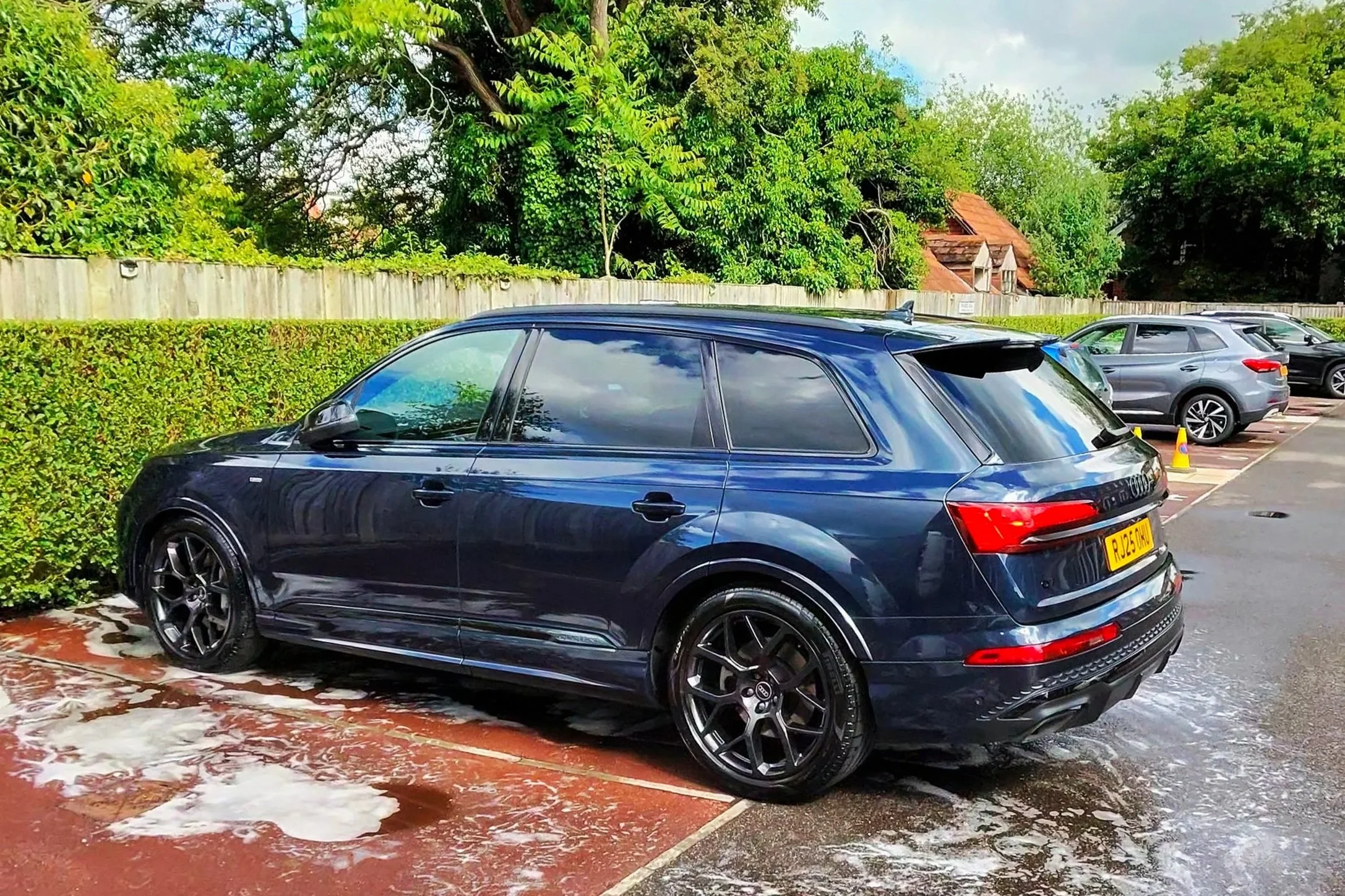 A black Audi SUV parked in a parking lot with foam on the ground after a washing. Other cars are parked nearby, and a wooden fence with green trees and houses in the background.