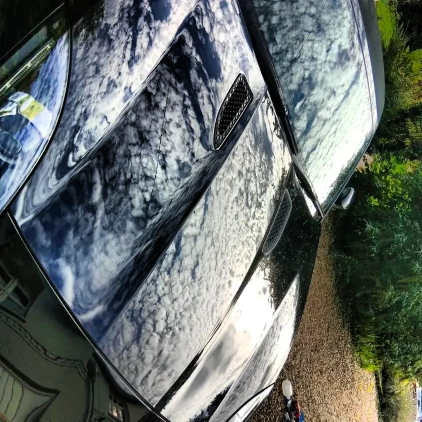 A black luxury car parked outdoors with reflections of clouds and sky on its shiny surface.