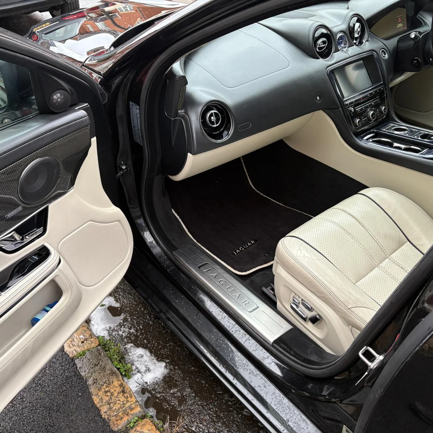 The interior of a black Jaguar vehicle showing the passenger side with beige leather seats, black dashboard, and floor mats with the Jaguar logo.