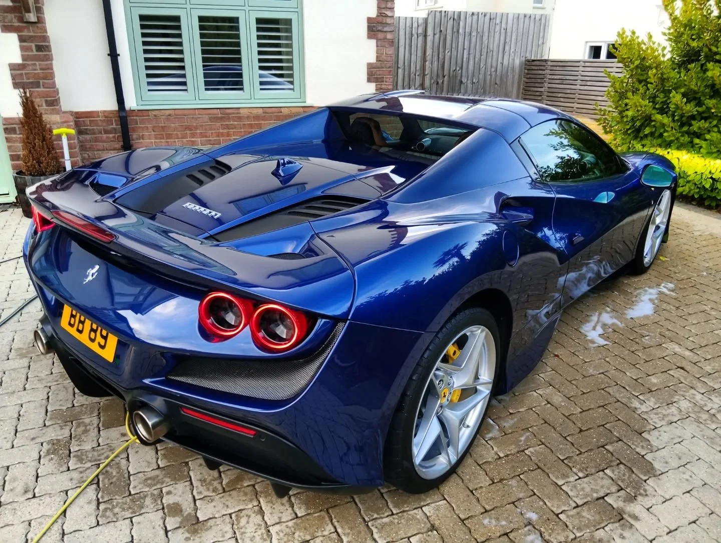 A blue Ferrari sports car parked on a brick driveway, with water around it and a green shrub on the right.