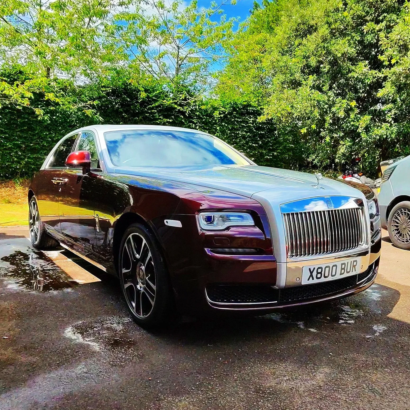 A black Rolls-Royce car with chrome grille and hood ornament parked outdoors on a wet surface with trees and blue sky in the background.