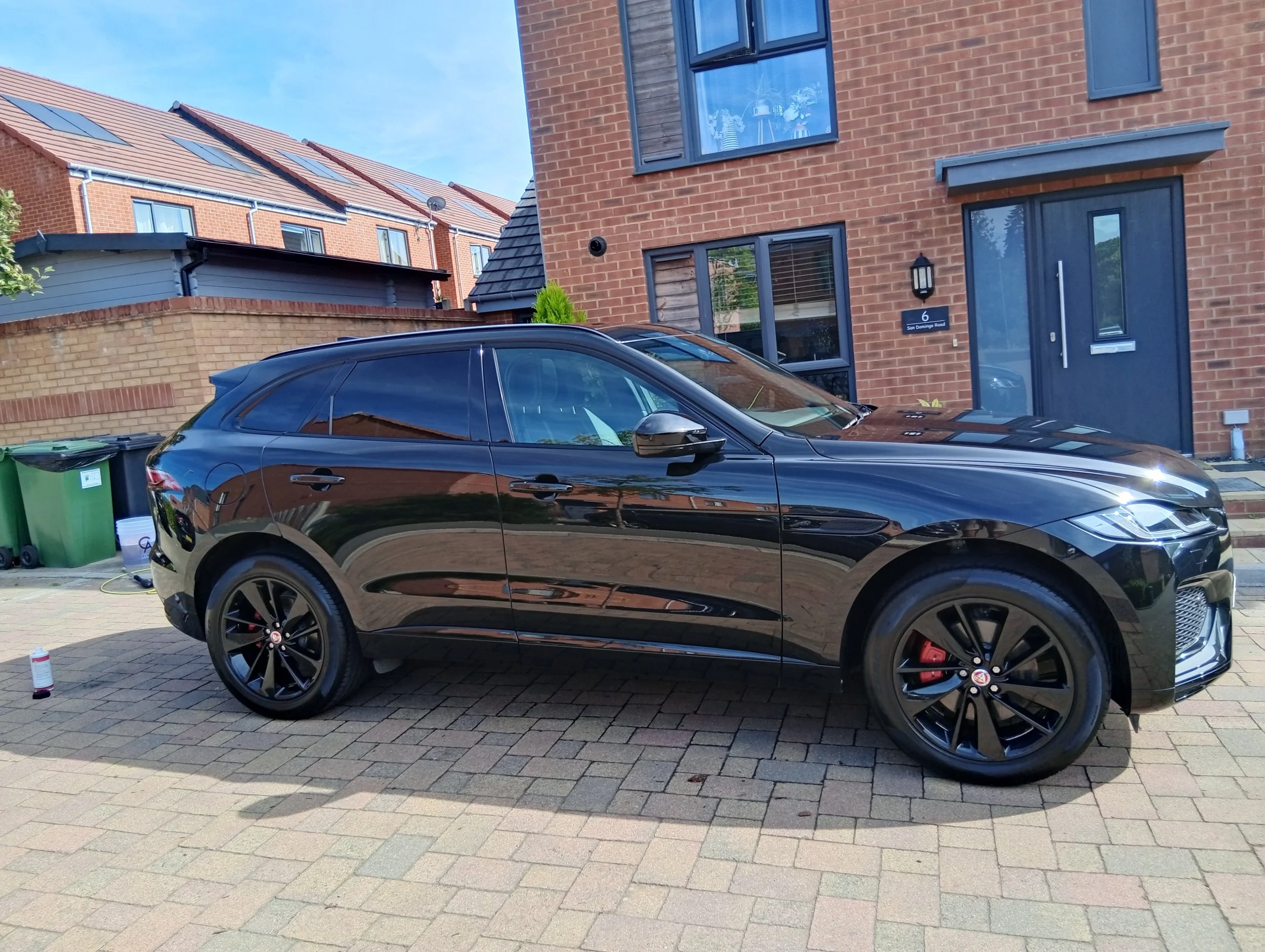 Black Jaguar parked on brick driveway in front of red brick residential building with blue door and windows, trash bins on the side, and a blue sky overhead.