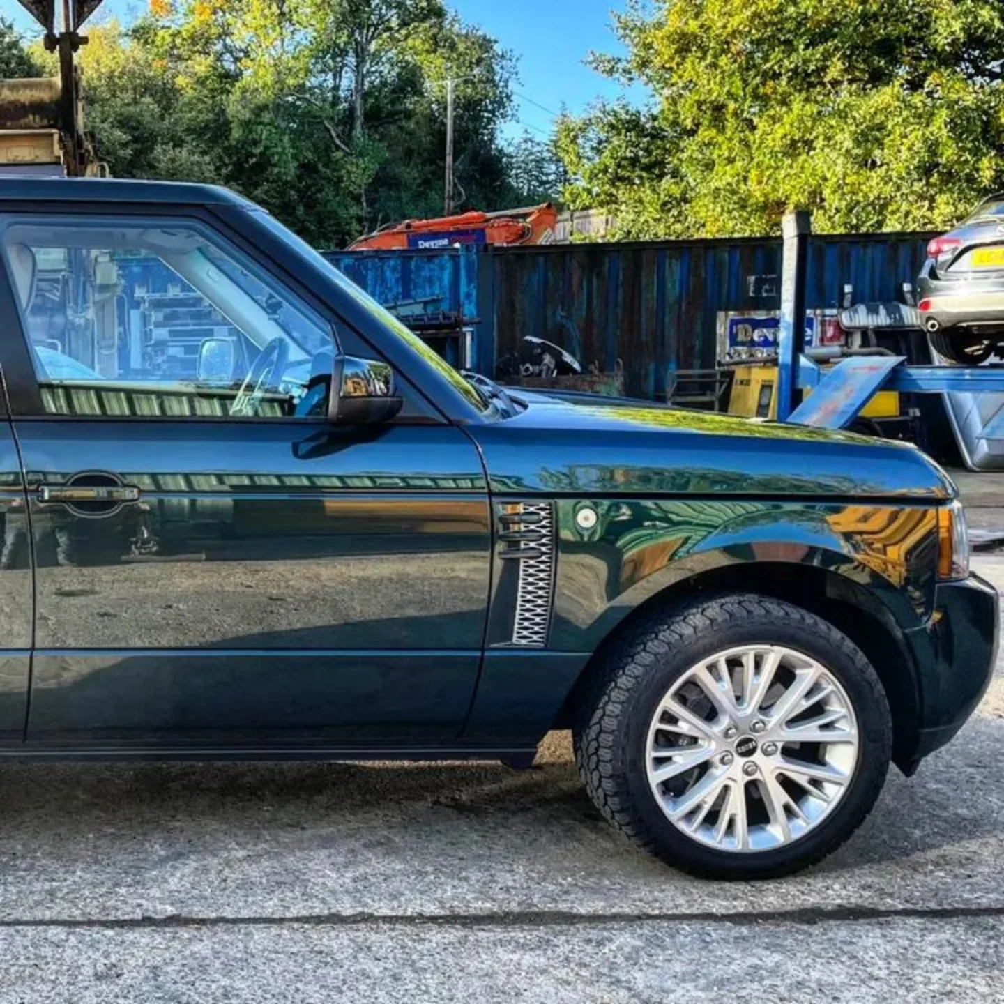 Close-up of a dark green Range Rover with the front half visible, parked outdoors on a gravel surface, with trees, a fence, and parked vehicles in the background.