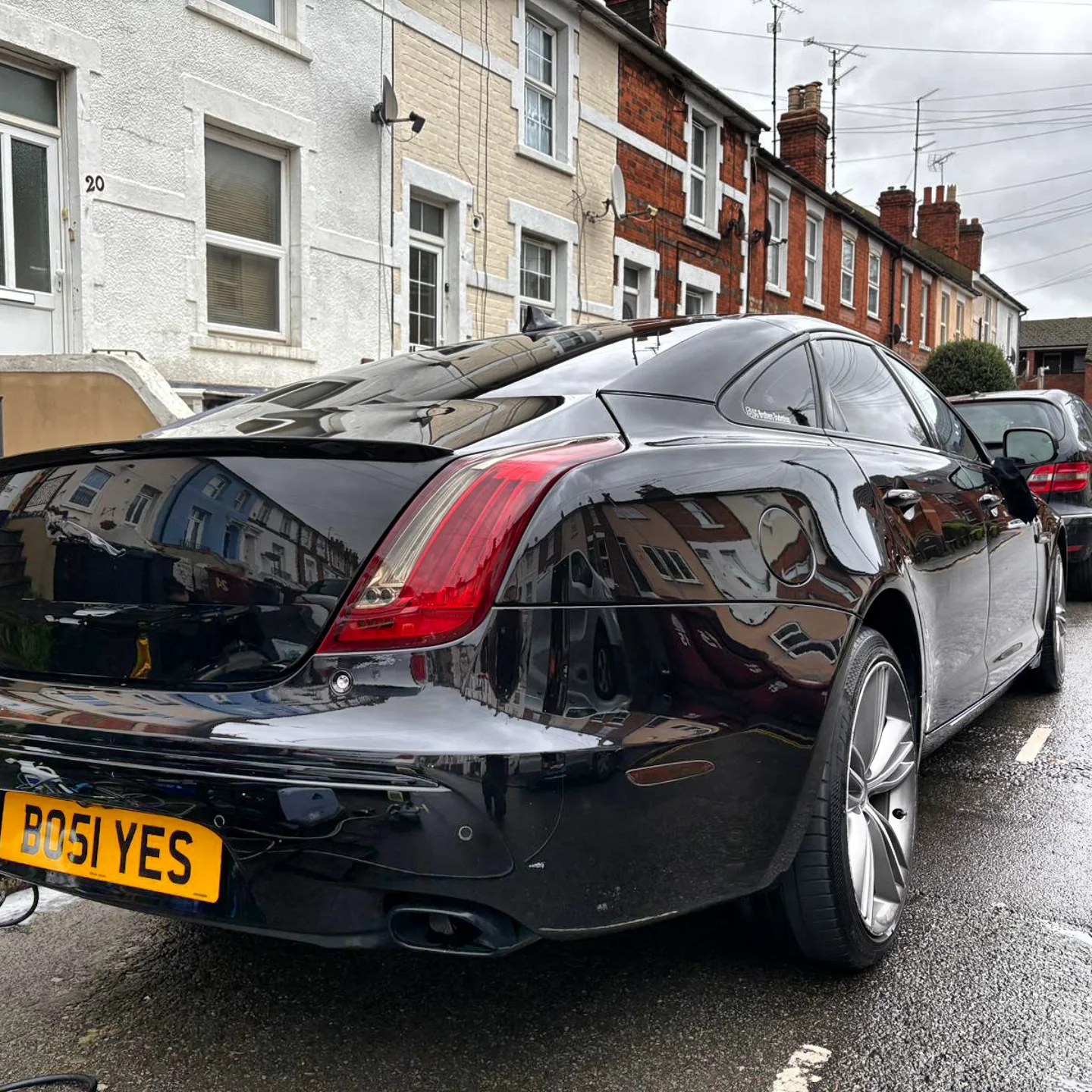 A black luxury sedan with a personalized yellow license plate that reads 'BOSI YES' parked on a residential street with brick and white houses in the background.