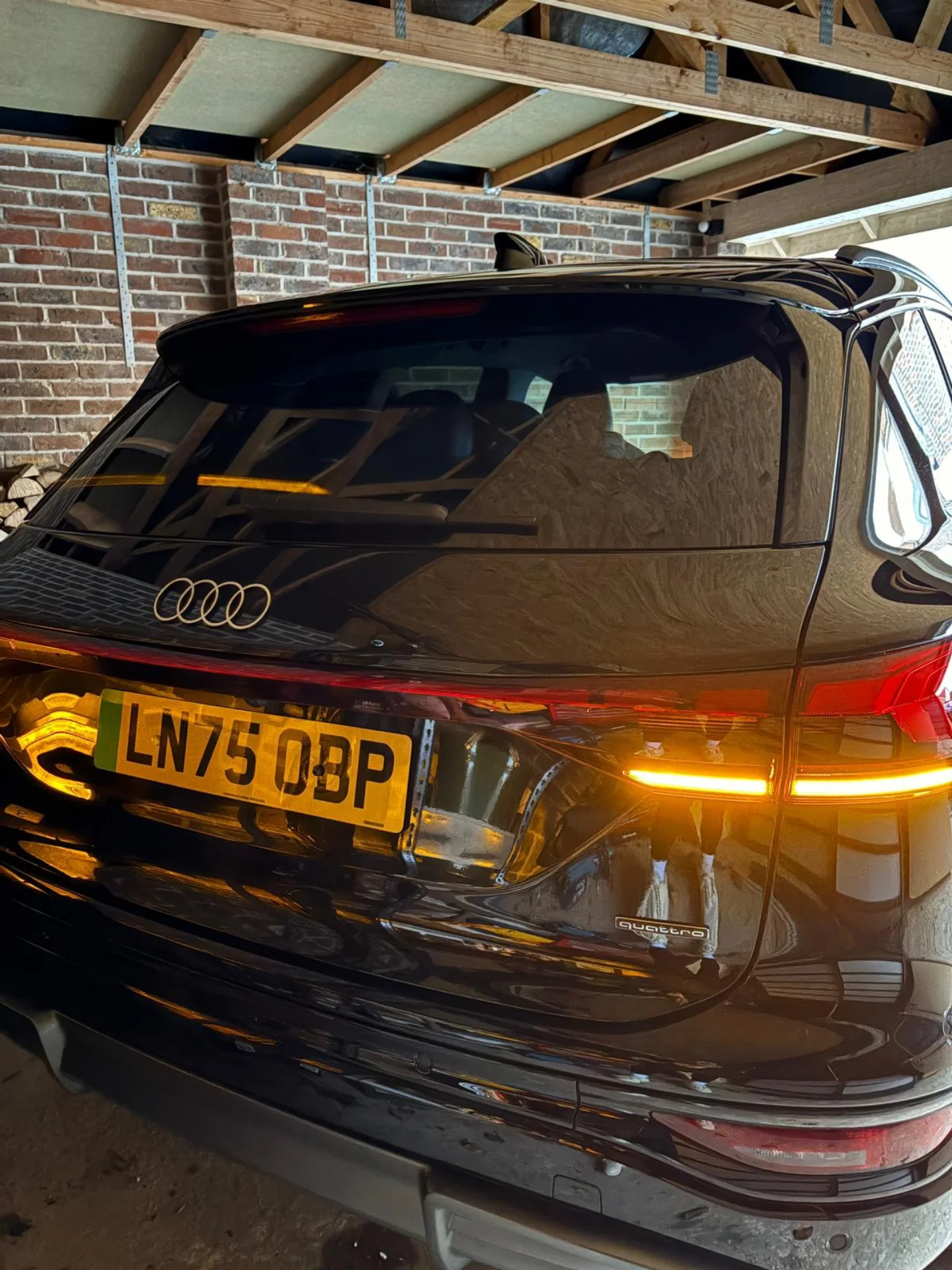Black Audi car parked in a garage with brick wall and wooden ceiling framing visible.