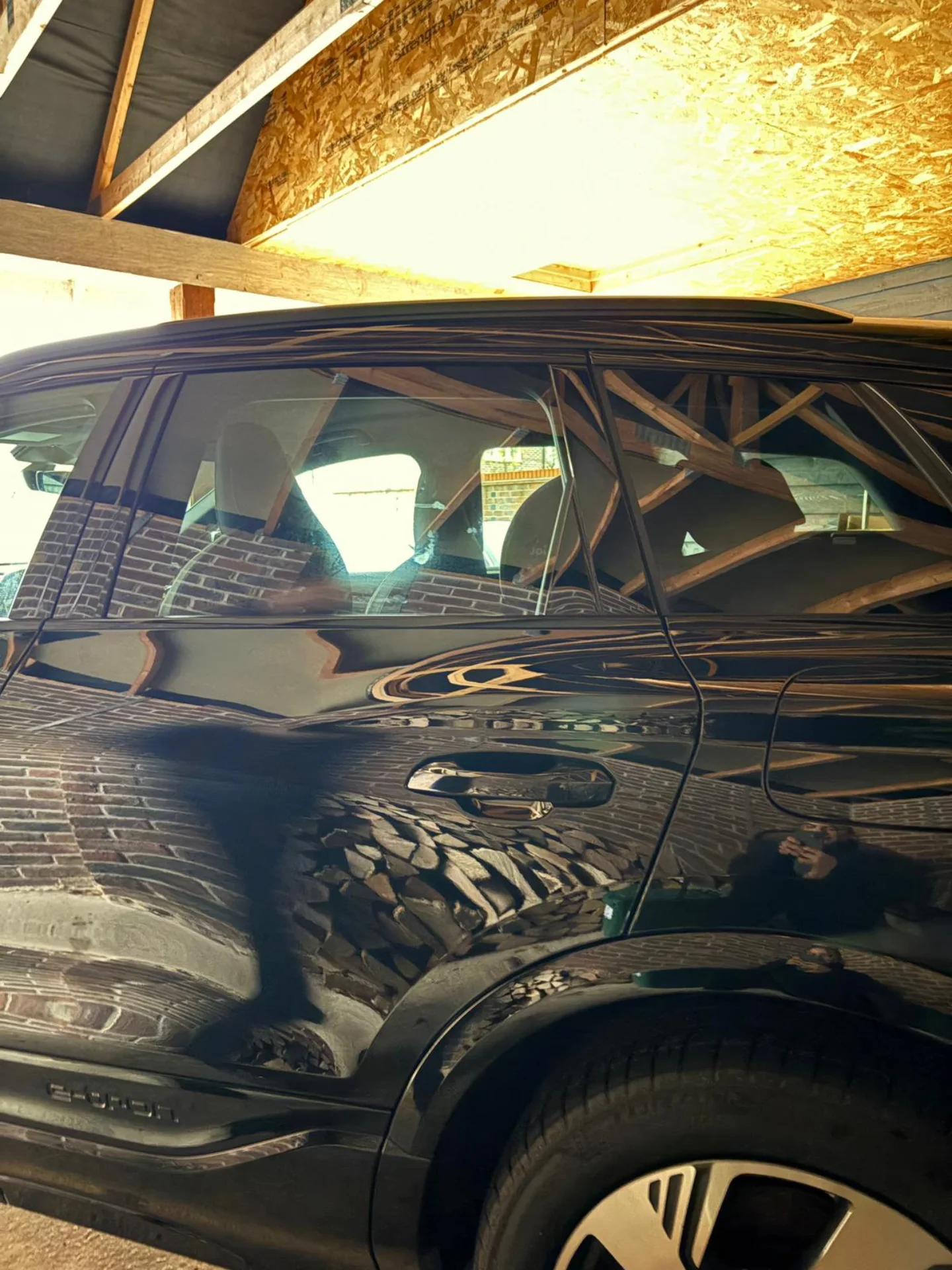 Black SUV parked inside a garage with brick wall and wooden ceiling. Reflection of a person taking the photo is visible on the car's surface.
