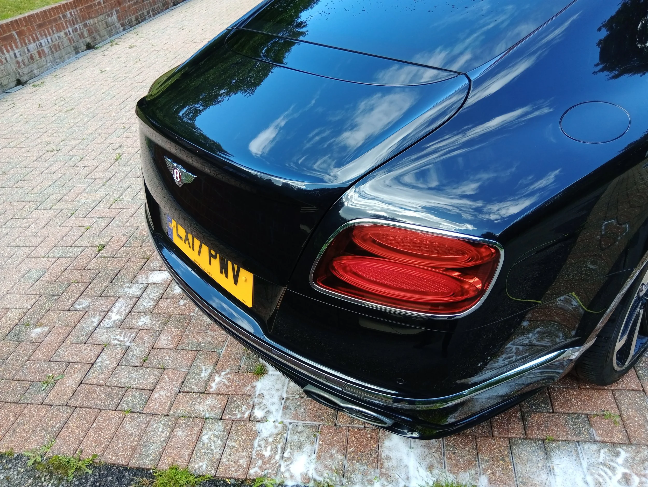 Close-up of the rear end of a shiny black Bentley car parked on a brick driveway, reflecting clouds and trees in the paint.