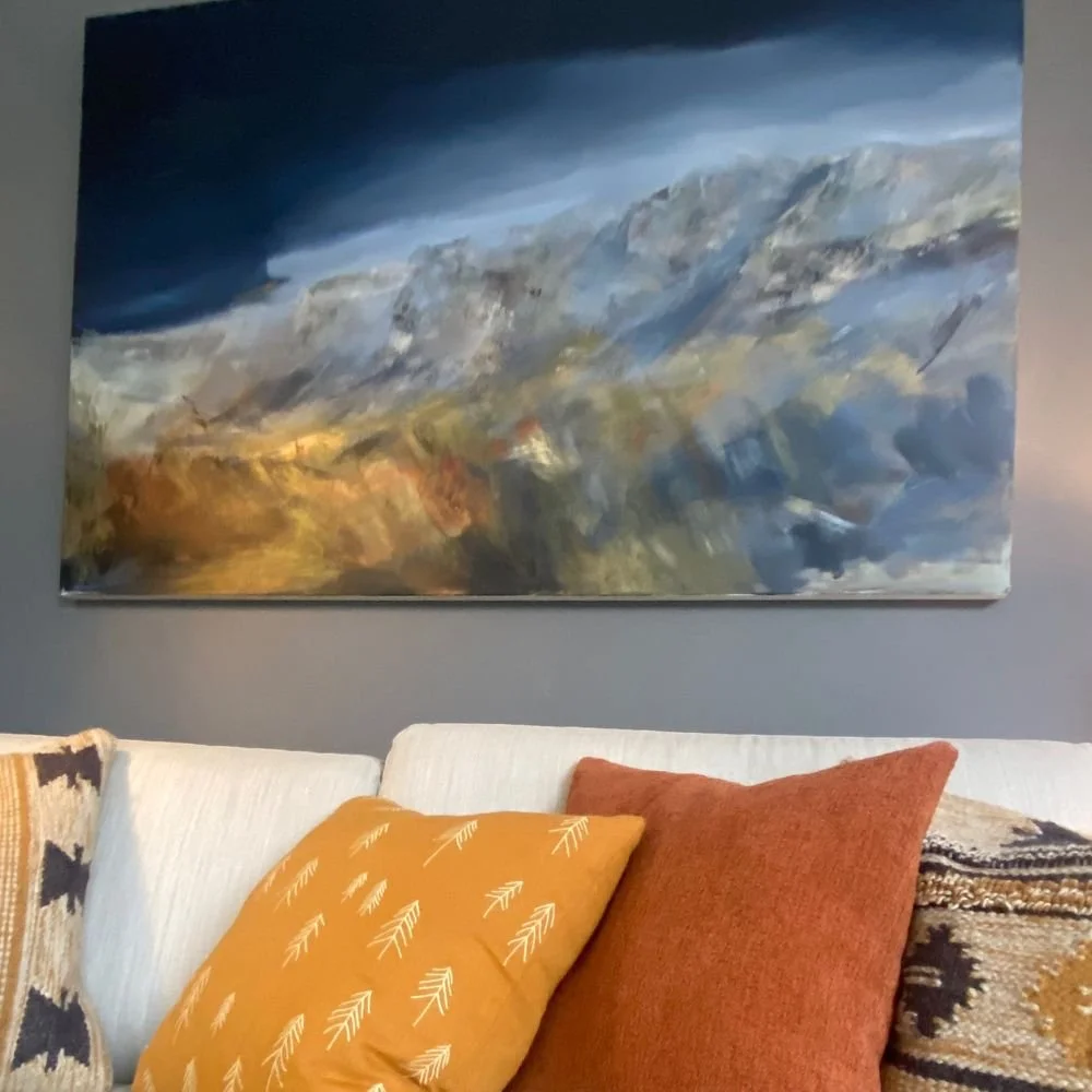 An abstract landscape painting of mountains with cloudy sky, hung above a white sofa with orange, mustard, and patterned pillows.