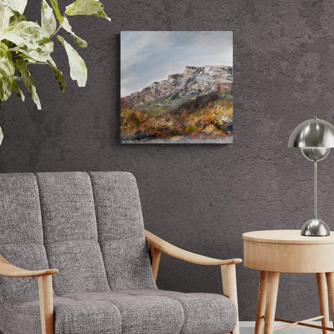 A modern living room with a gray upholstered armchair, a round wooden side table, a silver desk lamp, a green leafy plant in the upper left corner, and a landscape painting of mountains on a gray textured wall.