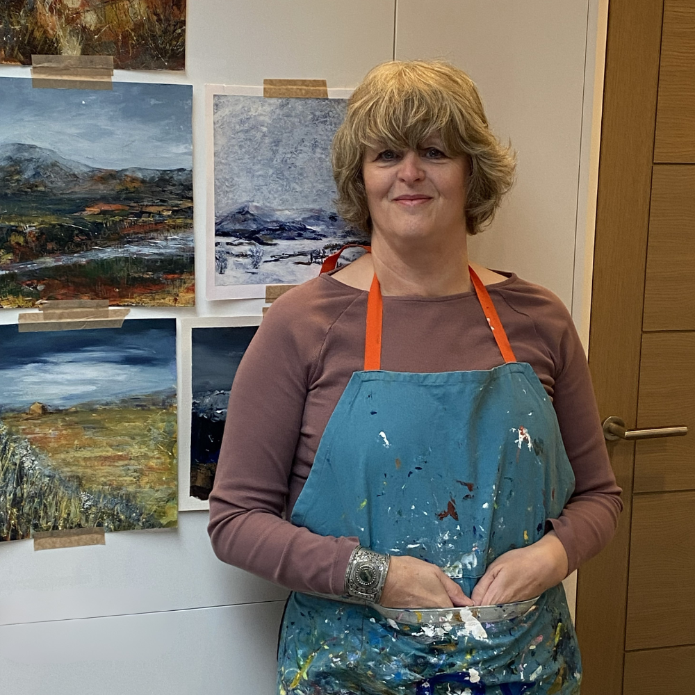 A woman holding a paint palette, standing in front of four landscape paintings taped to a wall.