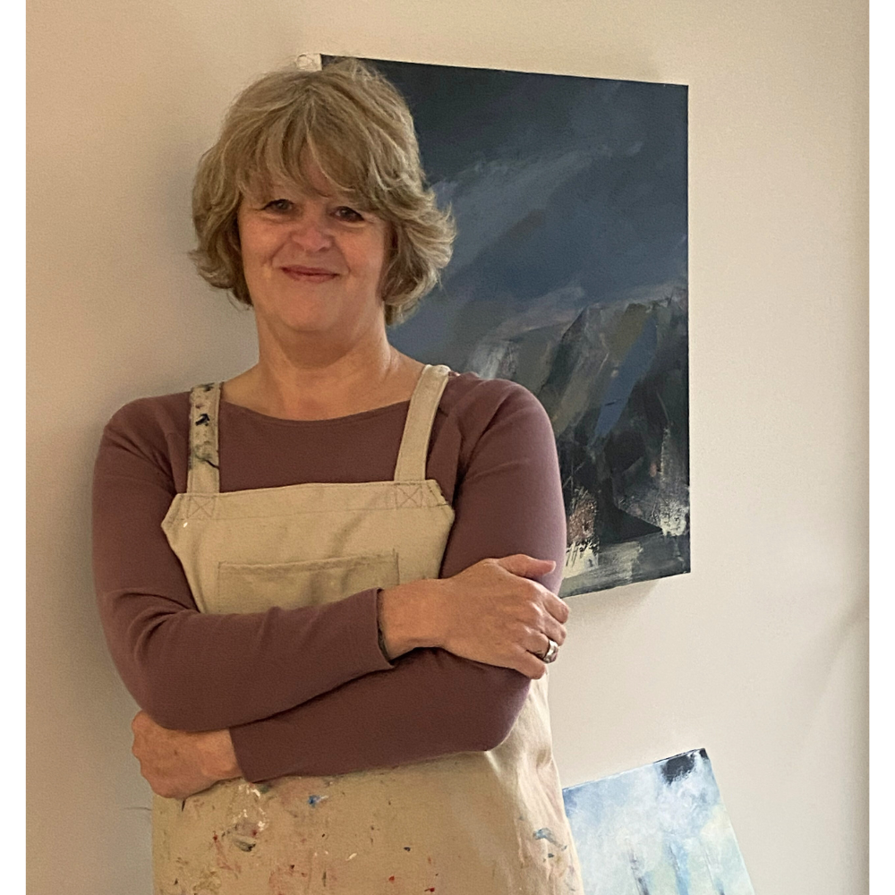 A Woman with short, wavy blonde hair standing with arms crossed, smiling, in front of a painting of abstract mountainous landscape, wearing a mauve long-sleeve shirt and a beige apron.