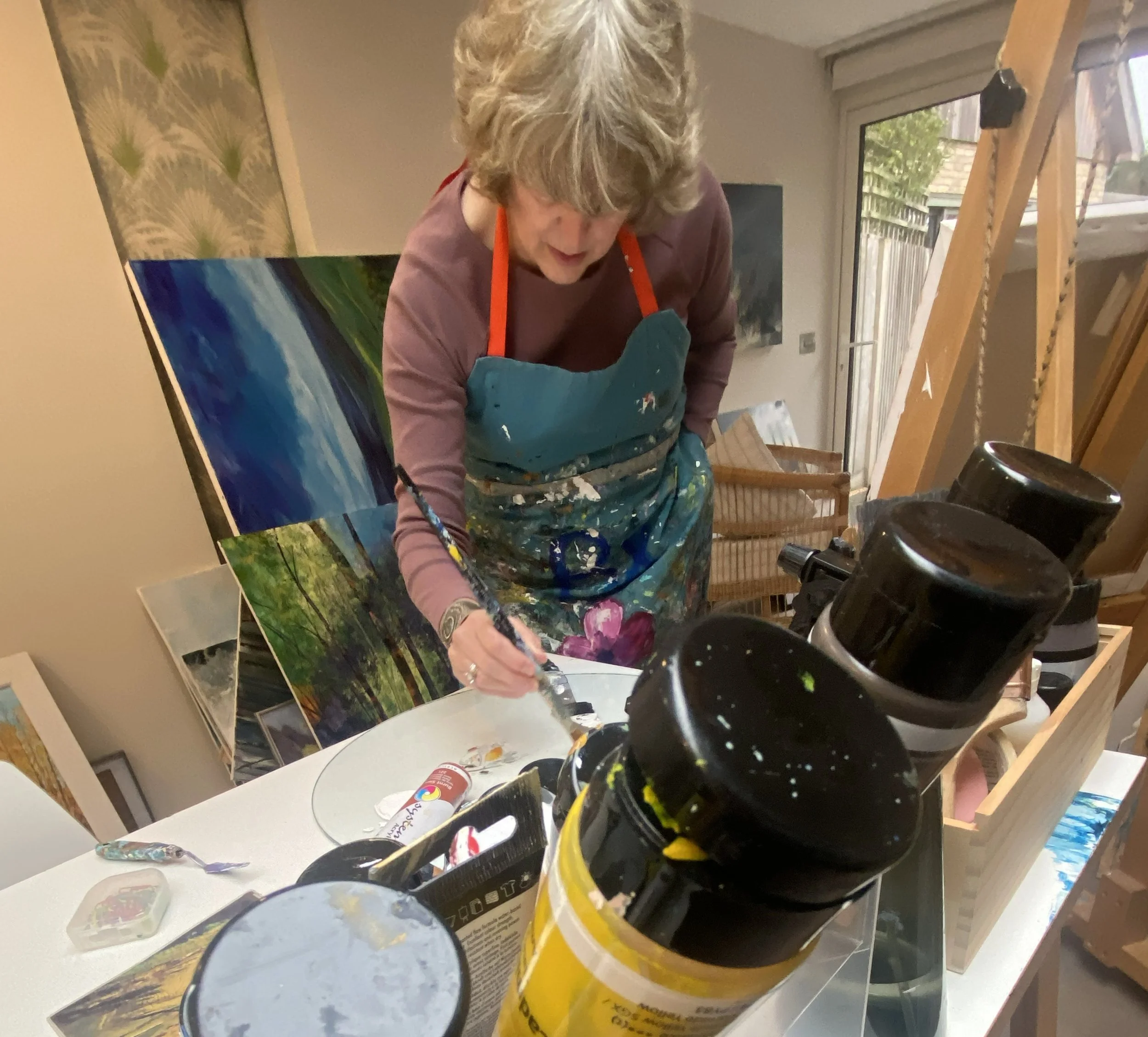 A woman with curly gray hair wearing a colorful apron, bending over a table, and painting outdoors in her studio. There are paintings, art supplies, and jars on the table, with a window and outdoor view in the background.