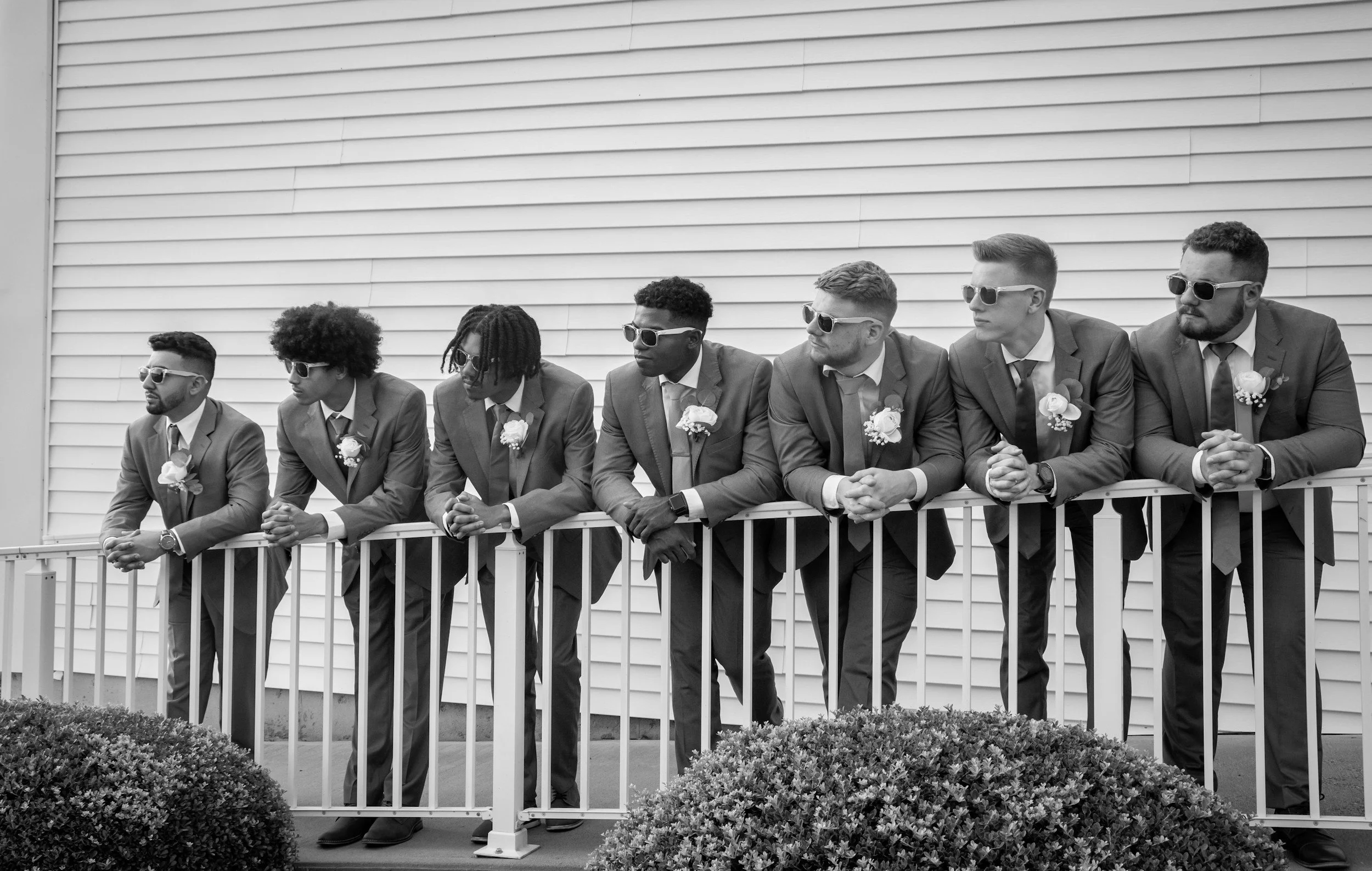 Seven men in suits standing behind a railing, wearing sunglasses and boutonnières, leaning on the railing with their arms crossed or resting on it, in front of a house with horizontal siding.