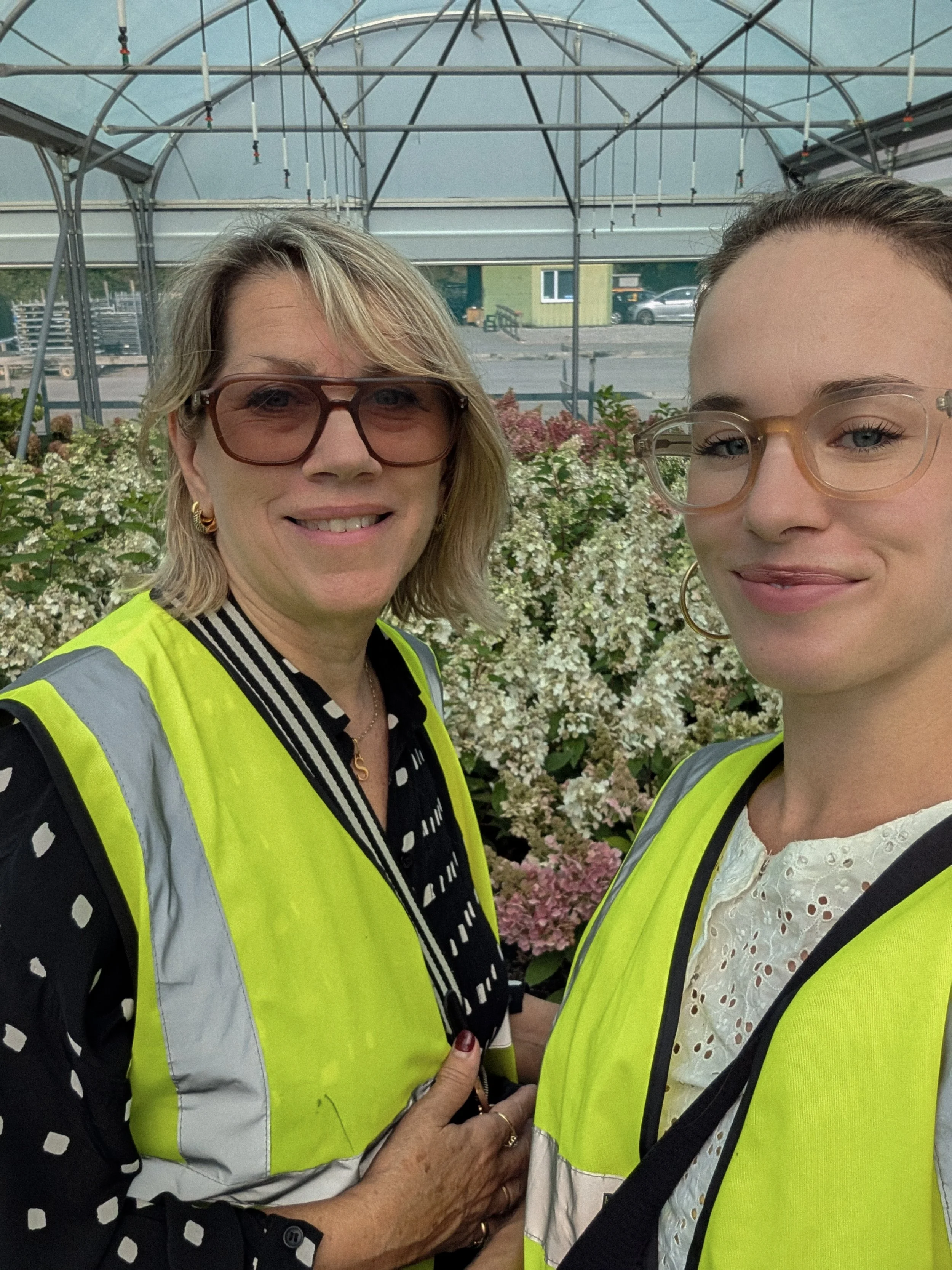 Two women smiling and holding hands, wearing yellow safety vests, standing in front of flowering bushes with a greenhouse structure in the background.