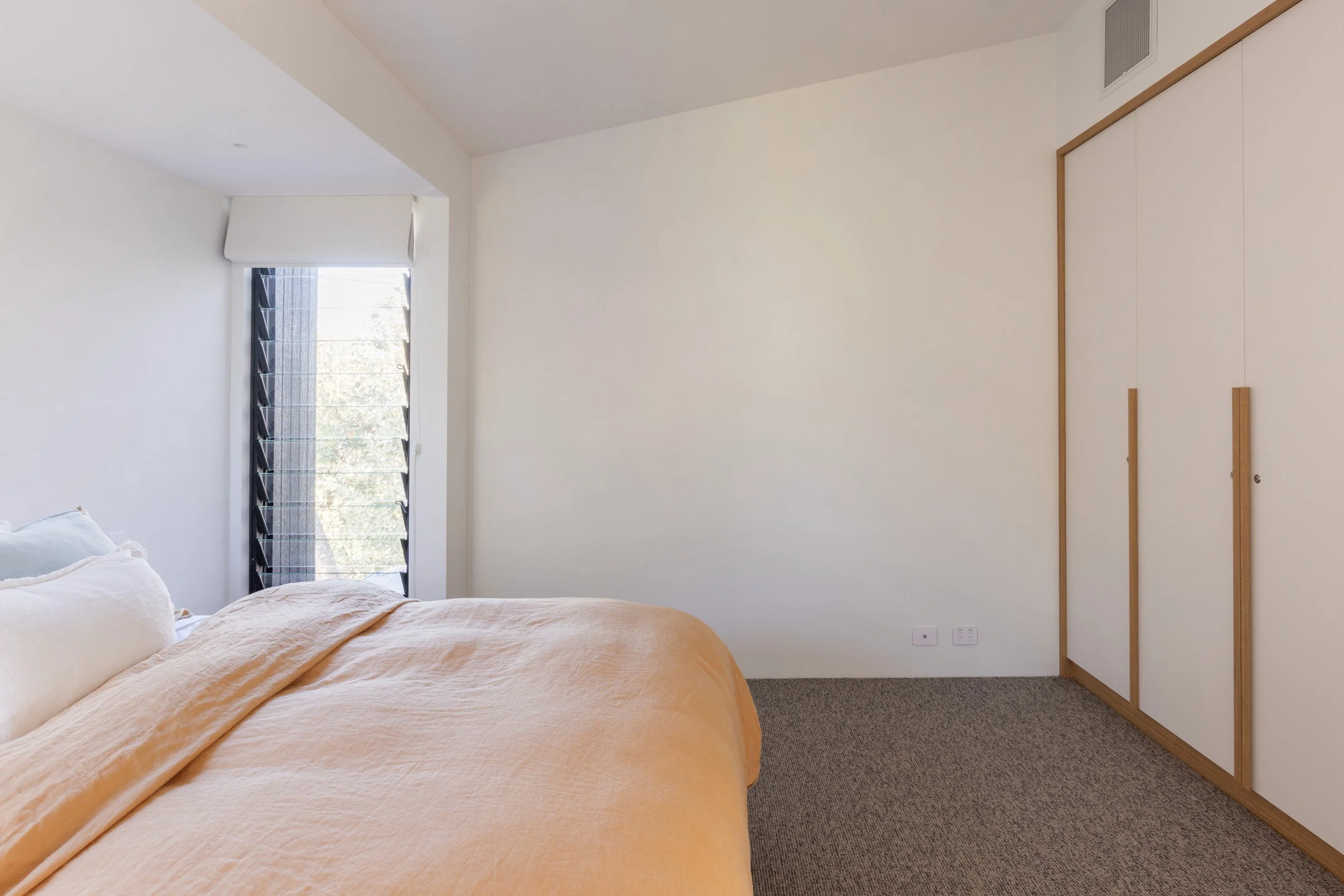 A corner of a bedroom with a bed covered with a peach-colored blanket, a window with blinds and a view outside, and a built-in wardrobe with wooden trim.