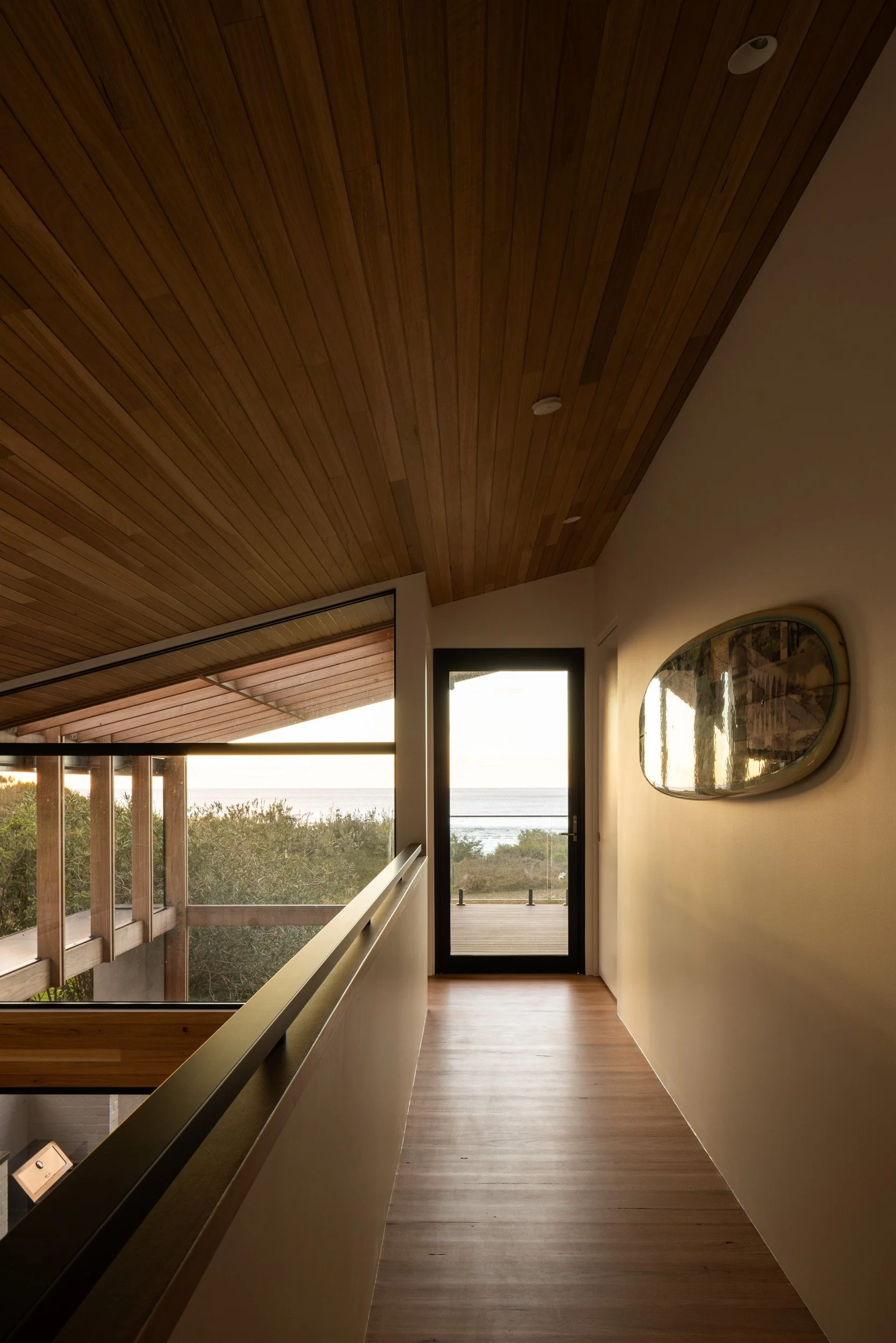 Interior view of a modern house hallway with wooden ceiling, beige walls, and a glass door leading outside to a deck facing the ocean during sunset.