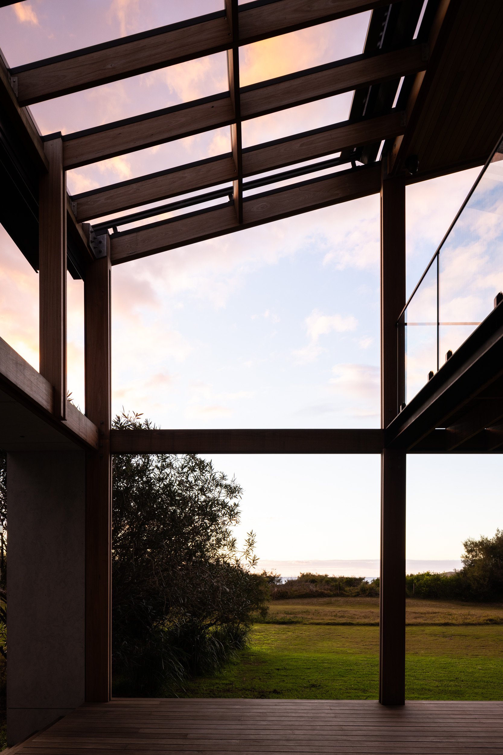 View of a modern wooden deck and open sky with pink clouds, trees, and grass beyond.