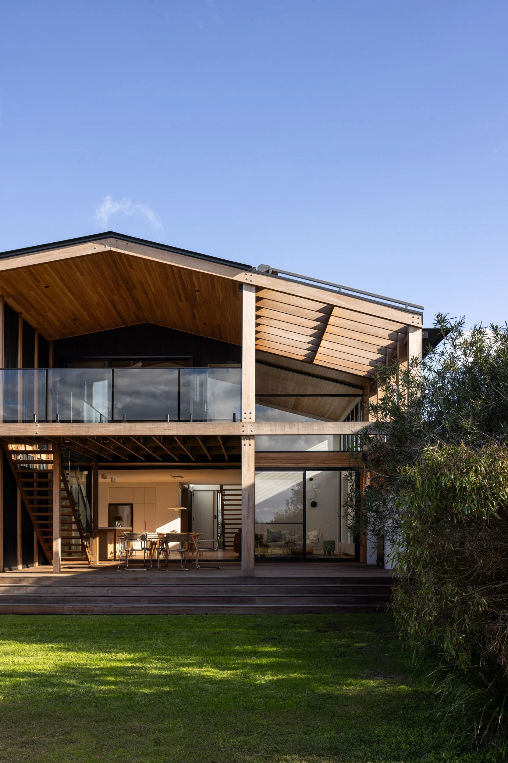 Modern two-story house with wooden exterior, glass balcony, and large sliding glass door, surrounded by green lawn and trees under a blue sky.