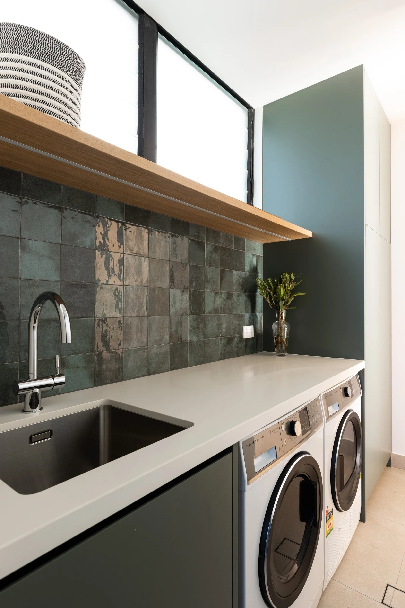 Laundry room with a white countertop, a sink, a green tiled backsplash, two front-loading washing machines, a vase with green leaves, and a wooden shelf above the sink.