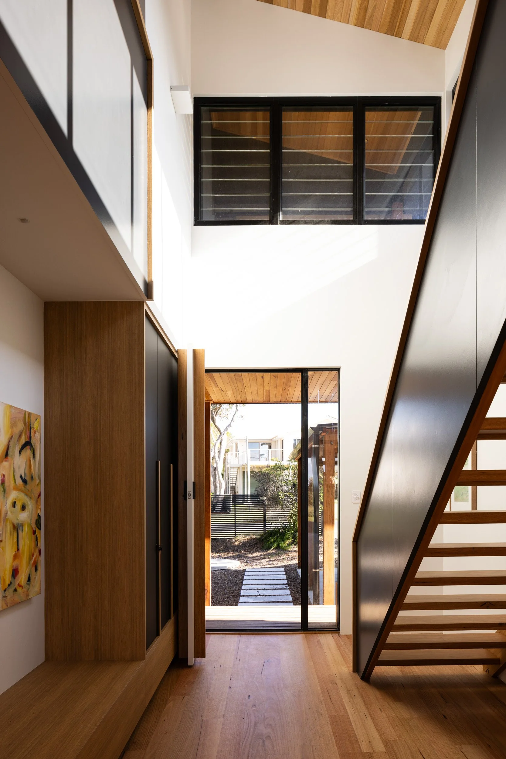 Interior view of a modern house entryway featuring wooden flooring, a staircase with black side paneling, a glass door leading outside, and large windows allowing natural light to fill the space.