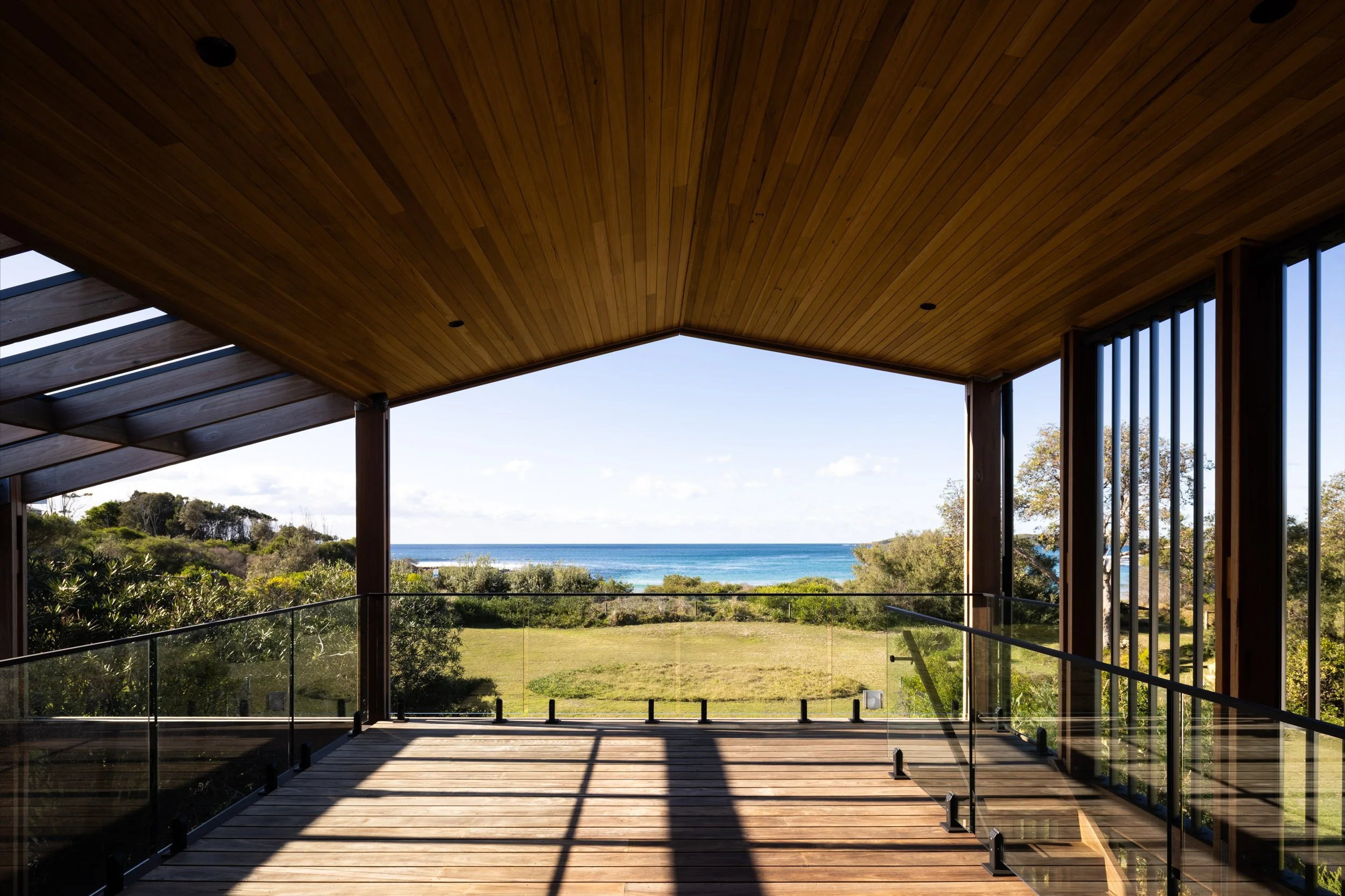 View from a wooden balcony overlooking green grass, trees, and the ocean under a partly cloudy sky.