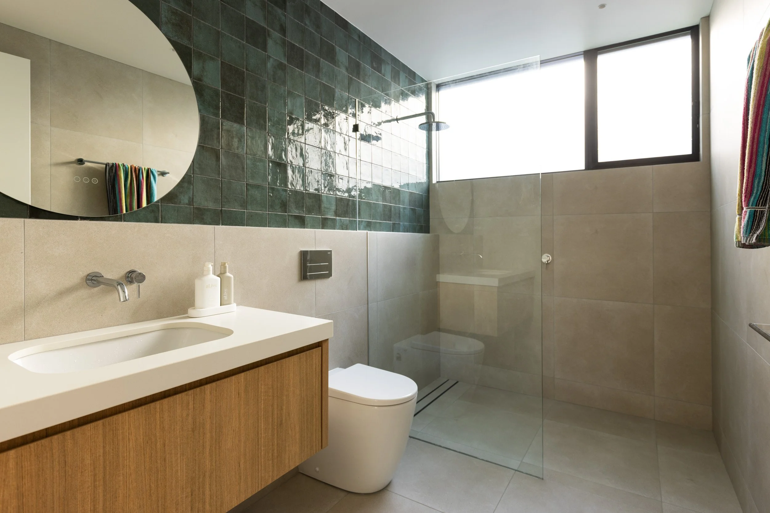 Modern bathroom with beige tiles, a floating wooden vanity with white countertop, a vessel-style sink, and a wall-mounted faucet. A round mirror hangs above the sink. There is a glass-enclosed shower with a large window, a rain showerhead, and a colo