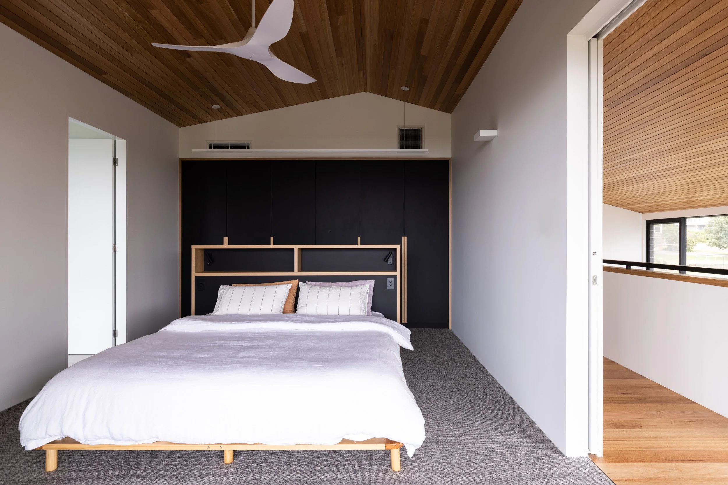 Modern bedroom with a bed featuring white bedding and pillows, black accent wall with wooden frame headboard, wood ceiling with white ceiling fan, and nearby white walls and sliding door open to a hallway with wooden flooring and large window.