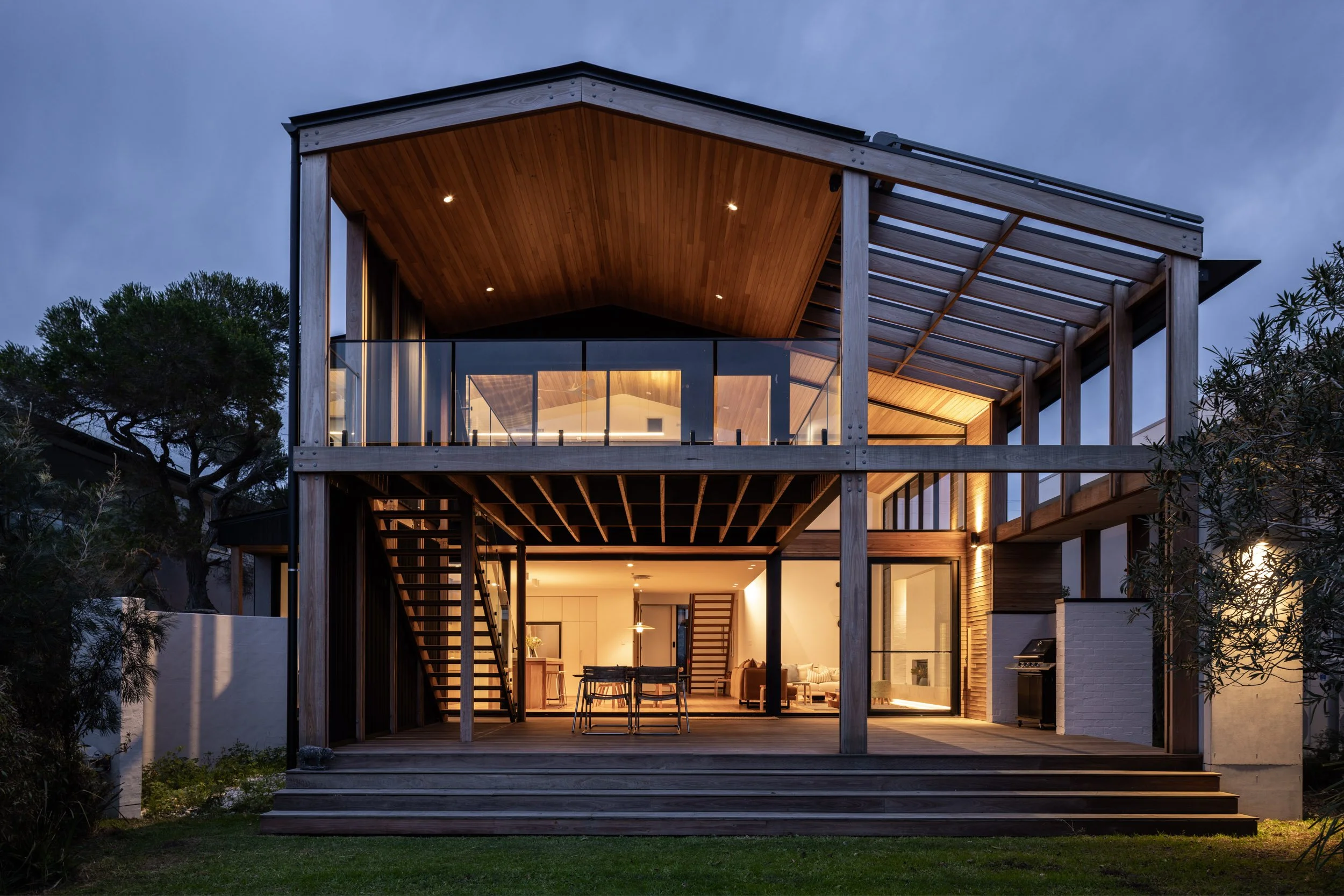 Modern two-story house with a wooden exterior, large glass windows, and an outdoor patio, illuminated at dusk.