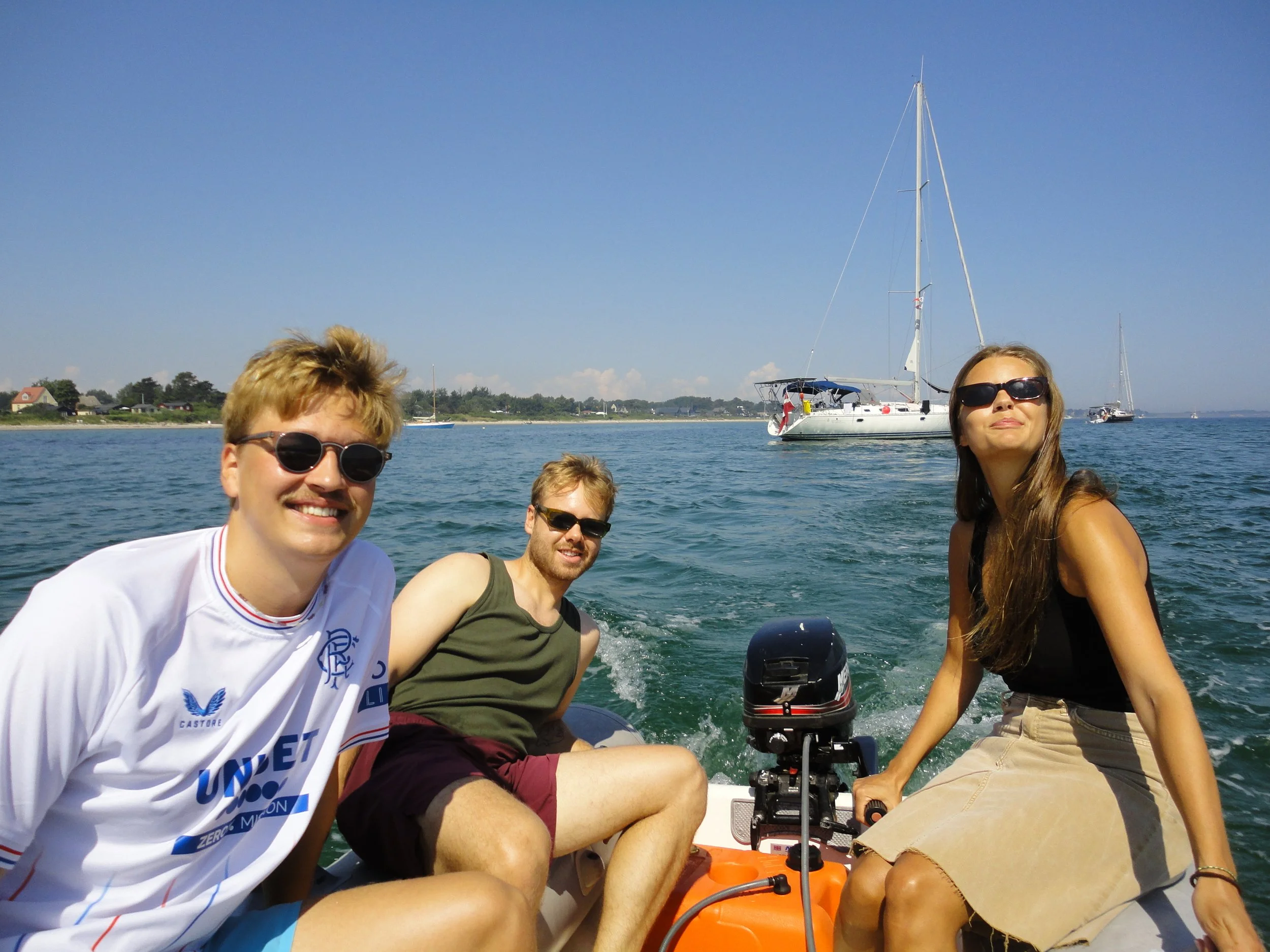 Three friends enjoying a boat ride on the water with sailboats in the background on a sunny day.