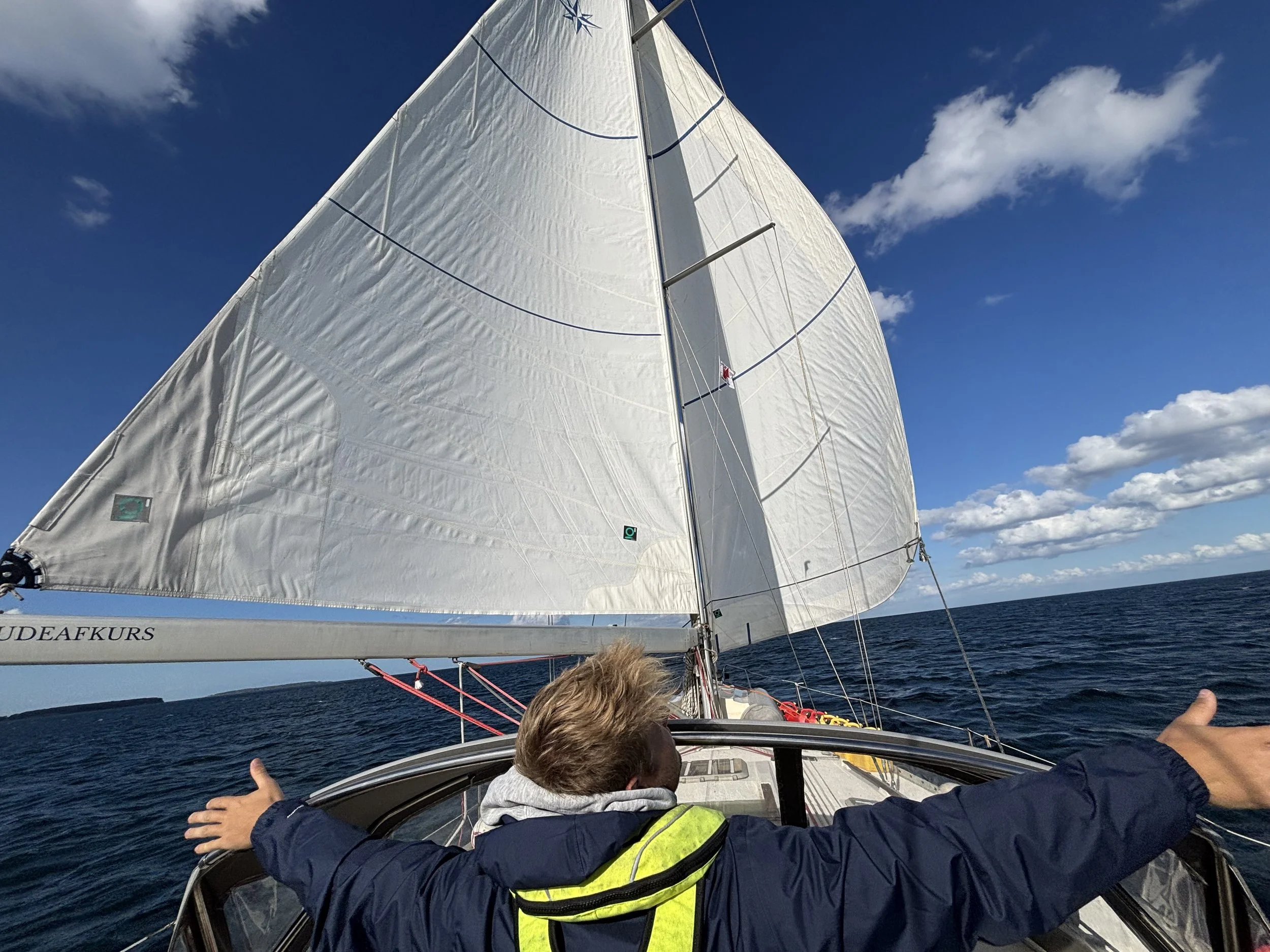 View from a sailboat of the ocean with a large white sail, blue sky with some clouds, and a person at the helm with arms outstretched.