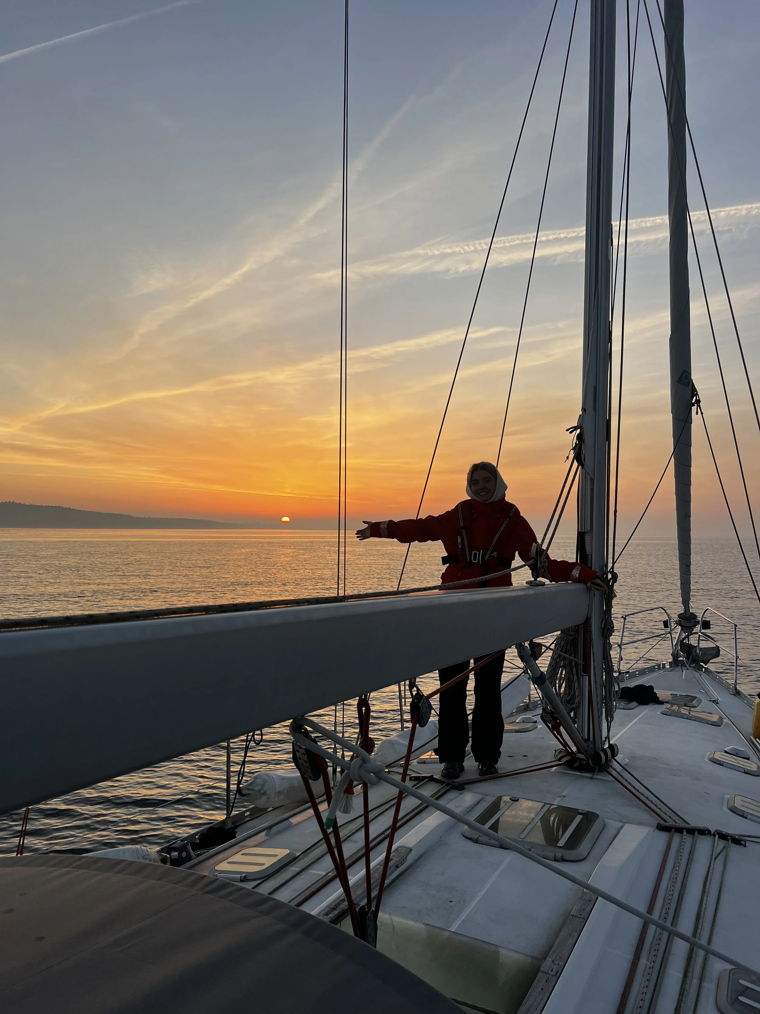 A person standing on a sailboat during sunset, smiling and reaching out with one arm, with the water and colorful sky in the background.
