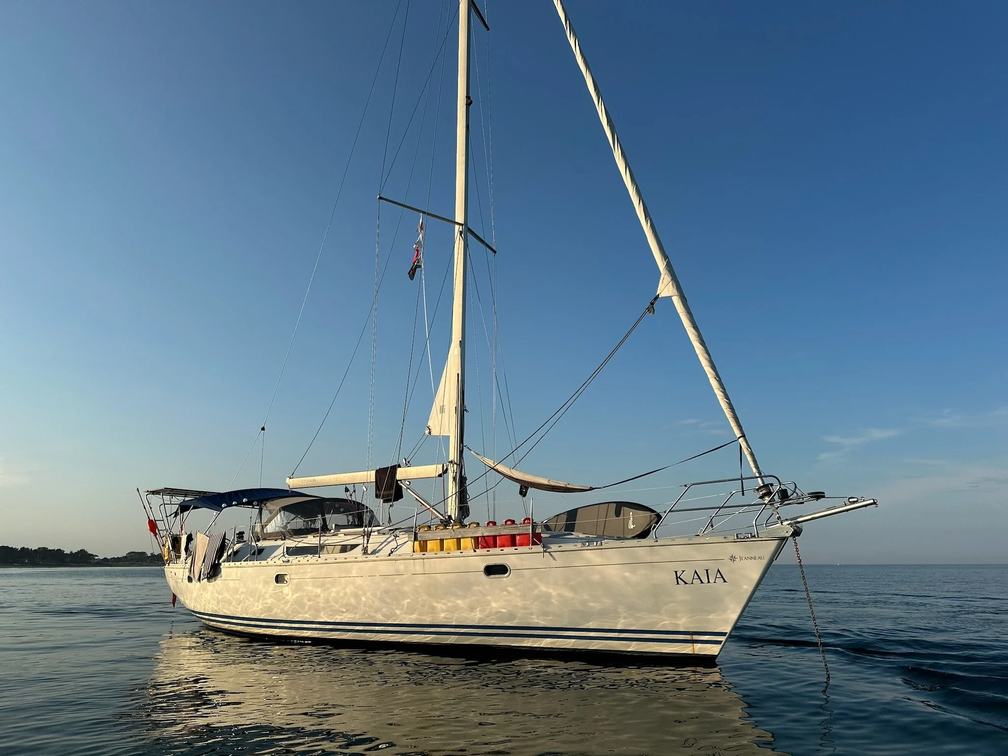 A white sailing yacht named Kaia floats on calm water near the coast during sunset, with a blue sky overhead.