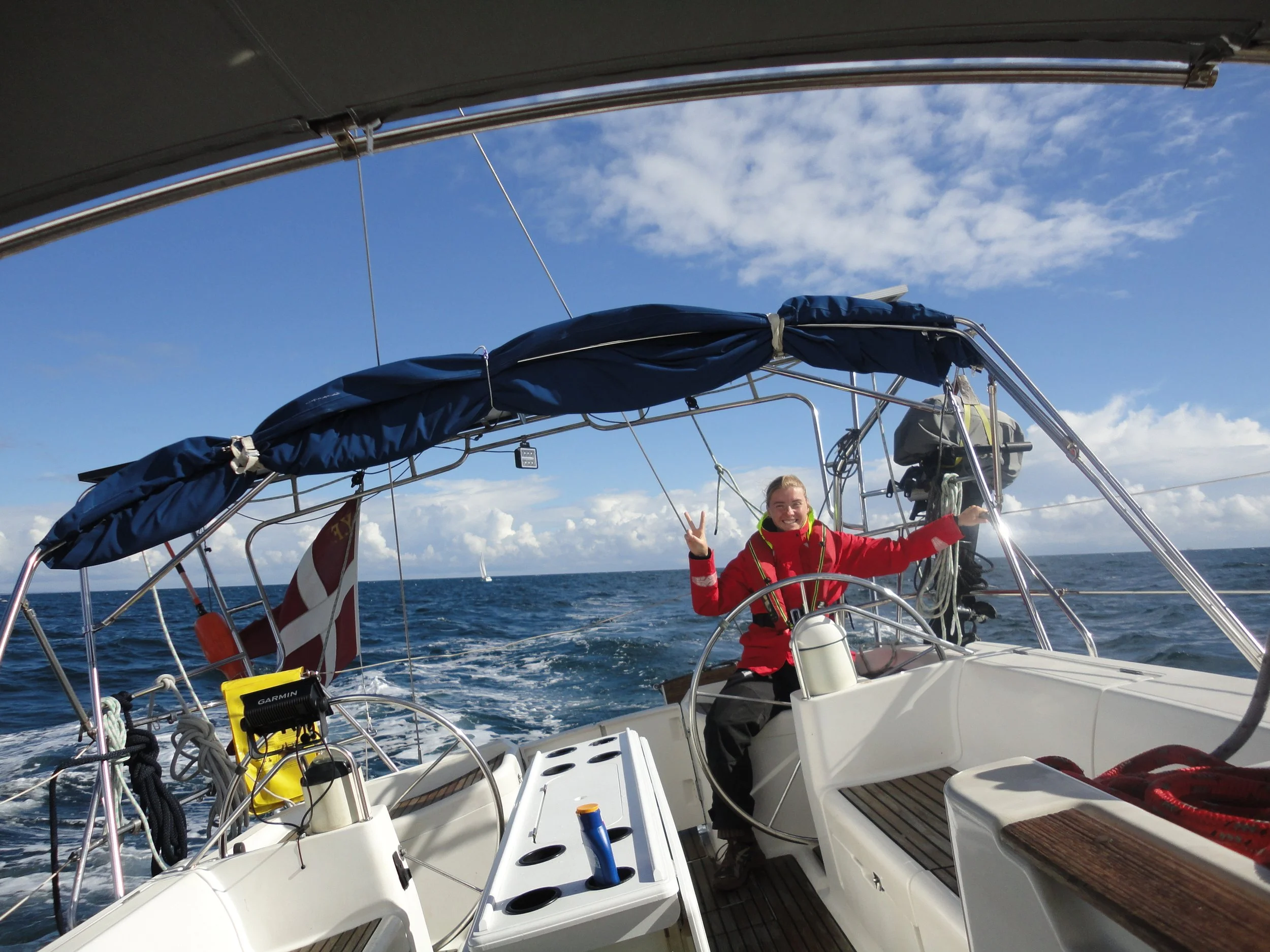 A woman on a sailboat smiling and making a peace sign, with blue sky and ocean in the background.