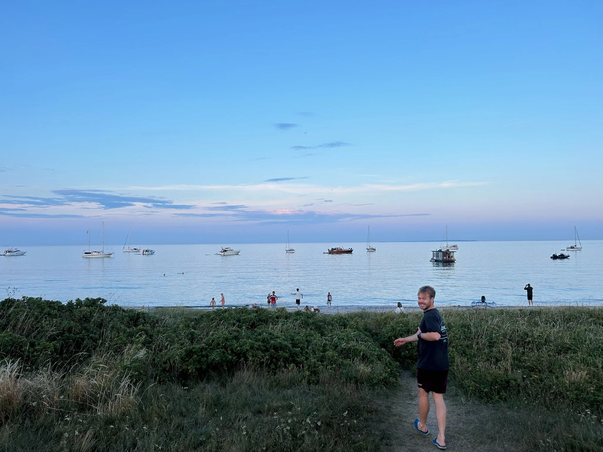 A man in a black shirt and shorts is walking along a grassy path near the beach, with a view of calm water, boats, and people on the beach during the evening sunset.