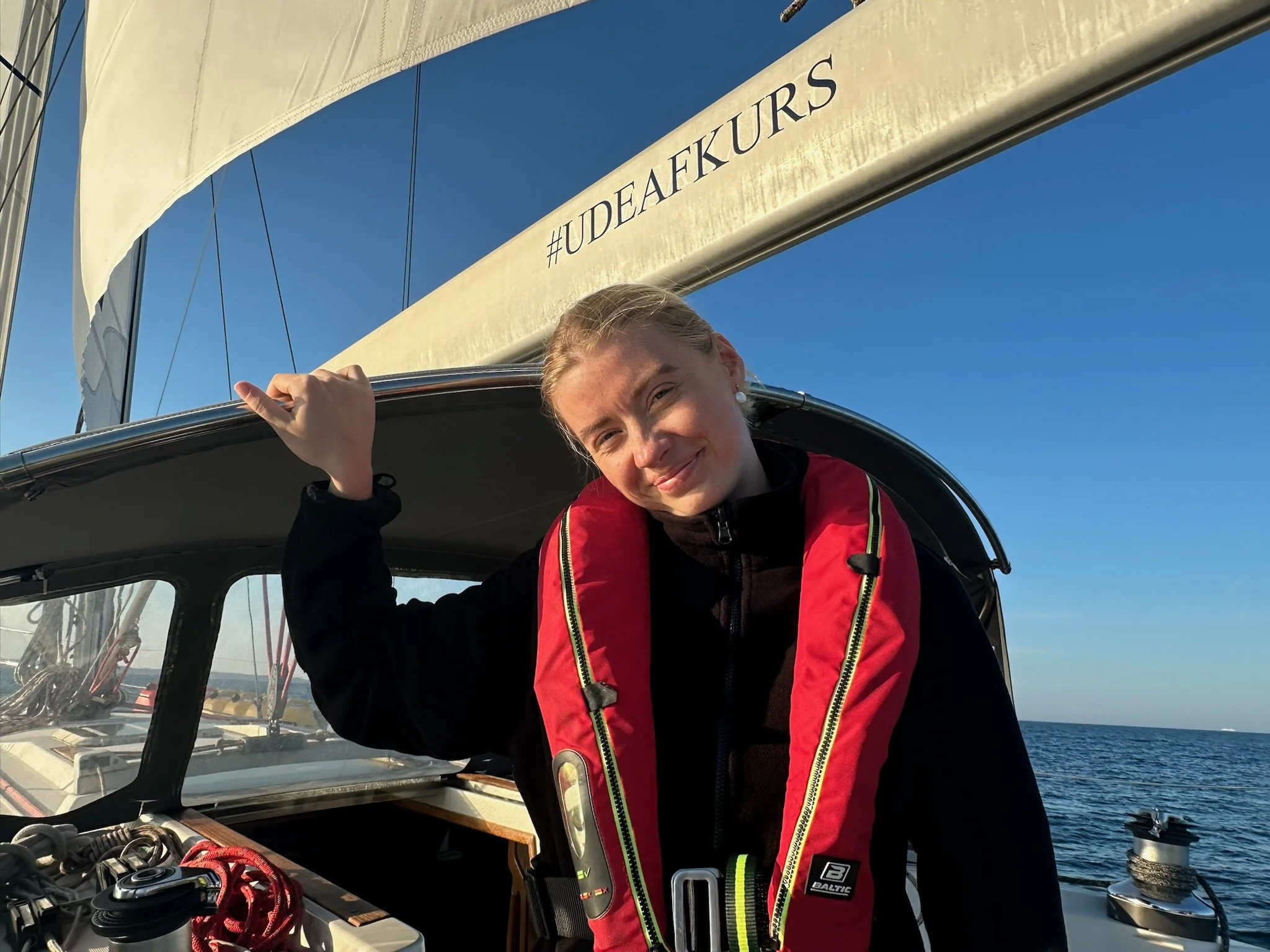 Young woman smiling, wearing a red life jacket, on a sailboat with blue sky and ocean in the background.